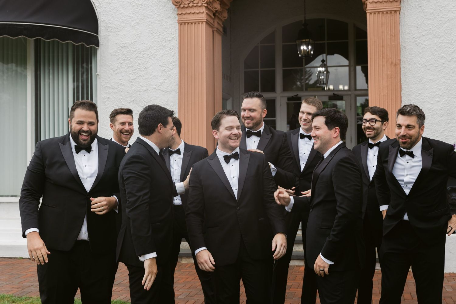 Groomsmen in black tuxedos check their phones while standing outside a venue.