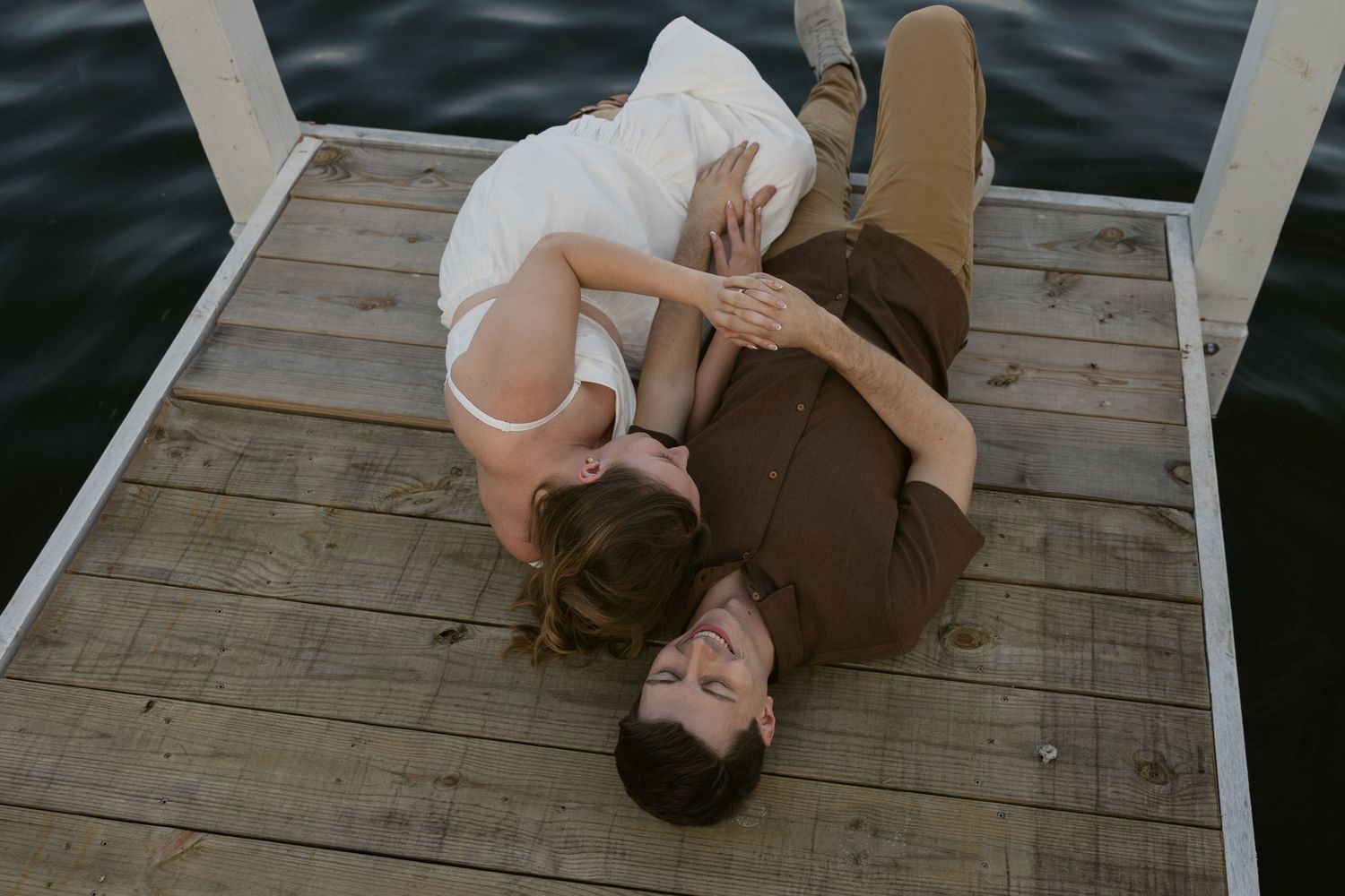 Two people laying together on a wooden dock over water during a romantic moment.