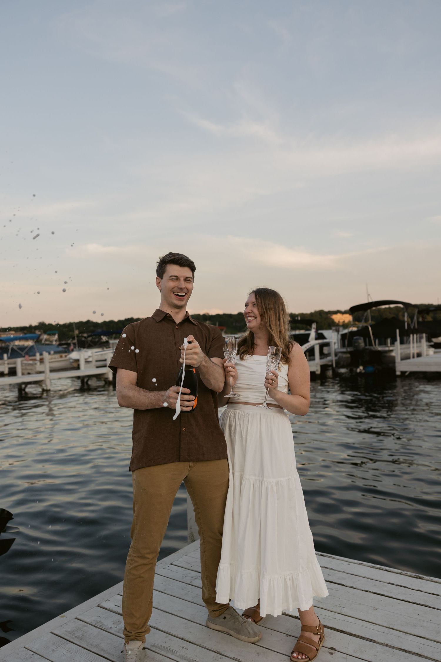 Couple laughing and embracing while standing on a wooden dock at sunset by the water.