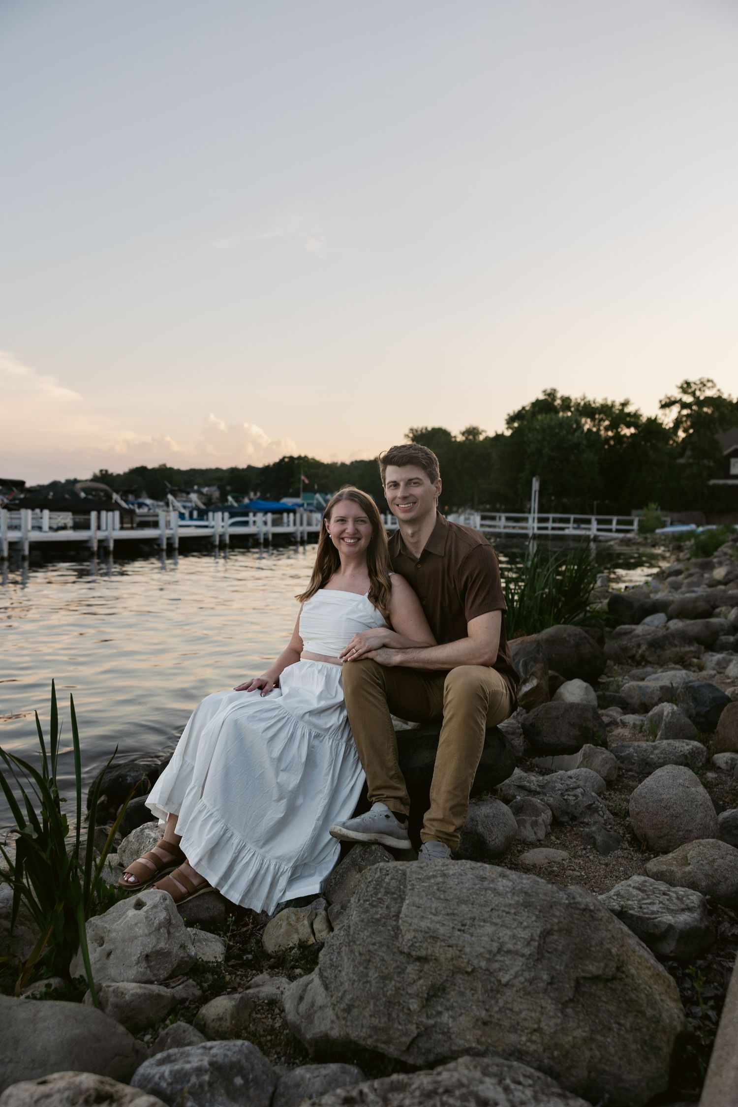 A romantic photo session on rocky shoreline with boats and water in background at dusk.