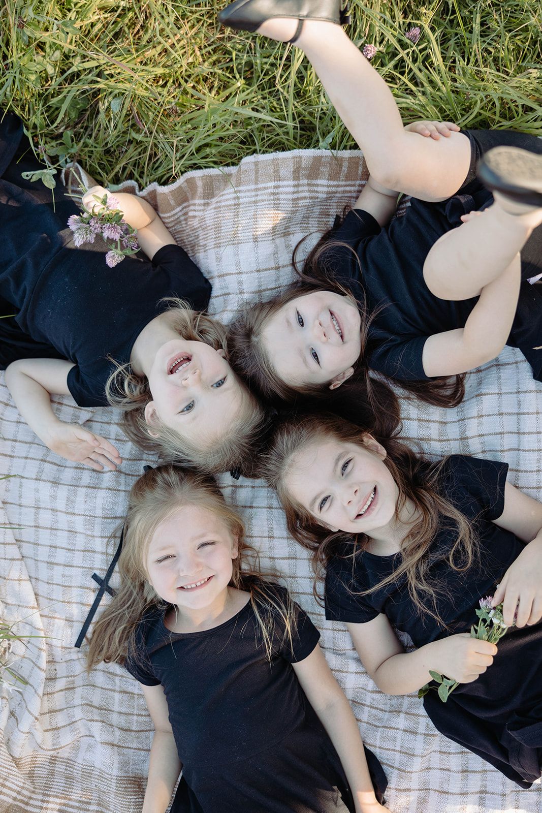 Four people lying on a picnic blanket in black outfits forming a circle while laughing outdoors.
