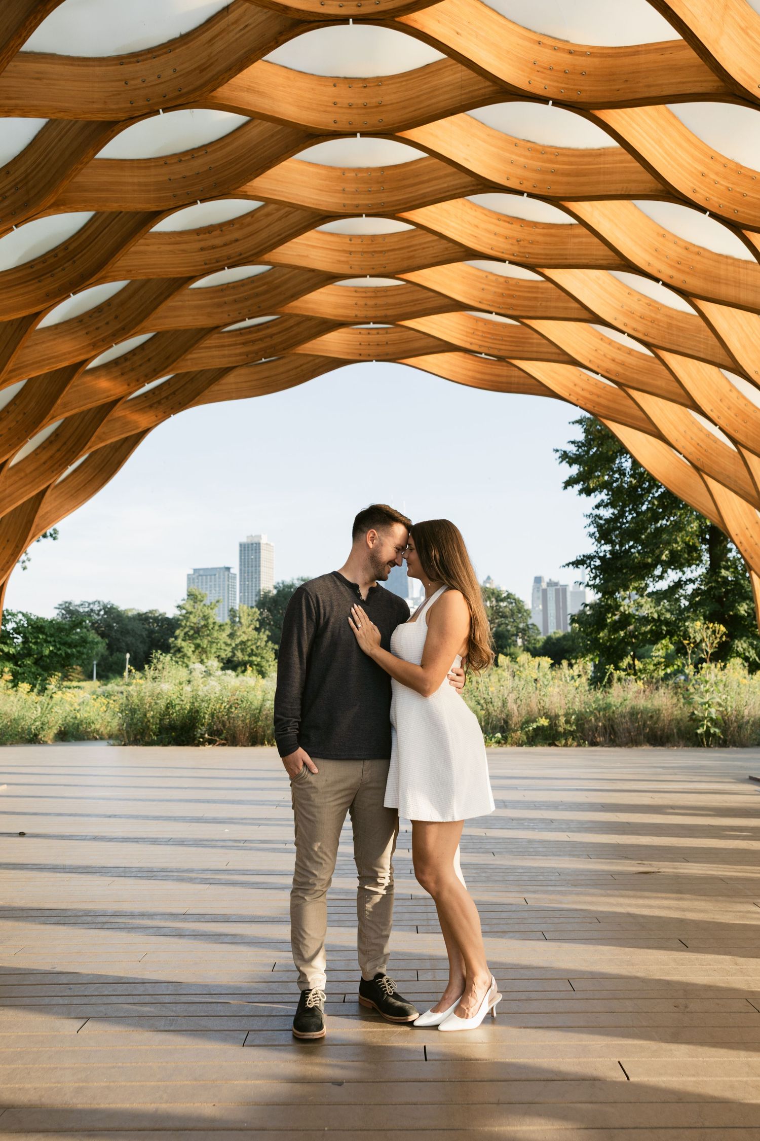 A couple shares a romantic moment under a curved wooden architectural structure with a city skyline visible in the background.