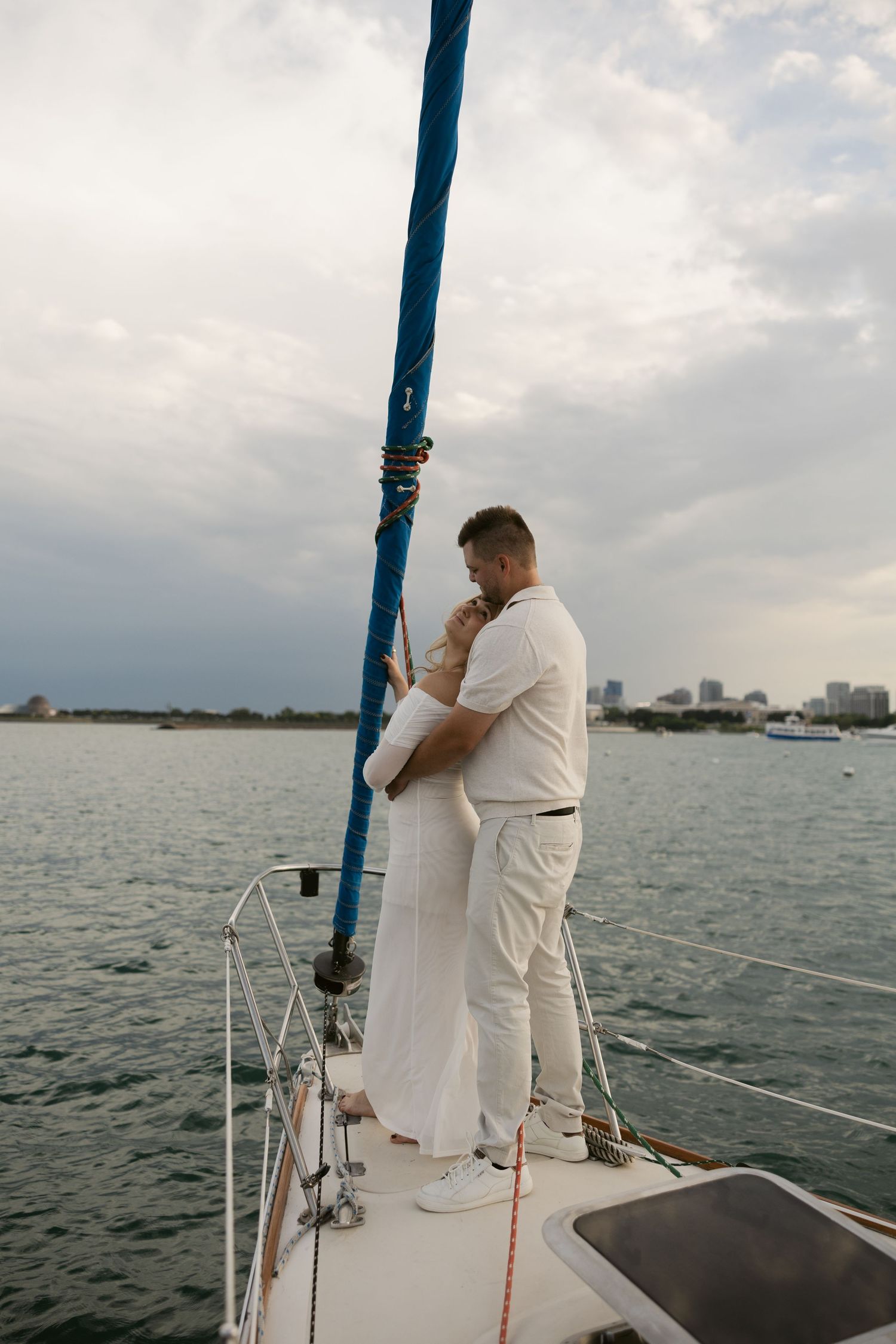 A couple embracing on the bow of a sailboat against a cloudy coastal skyline.