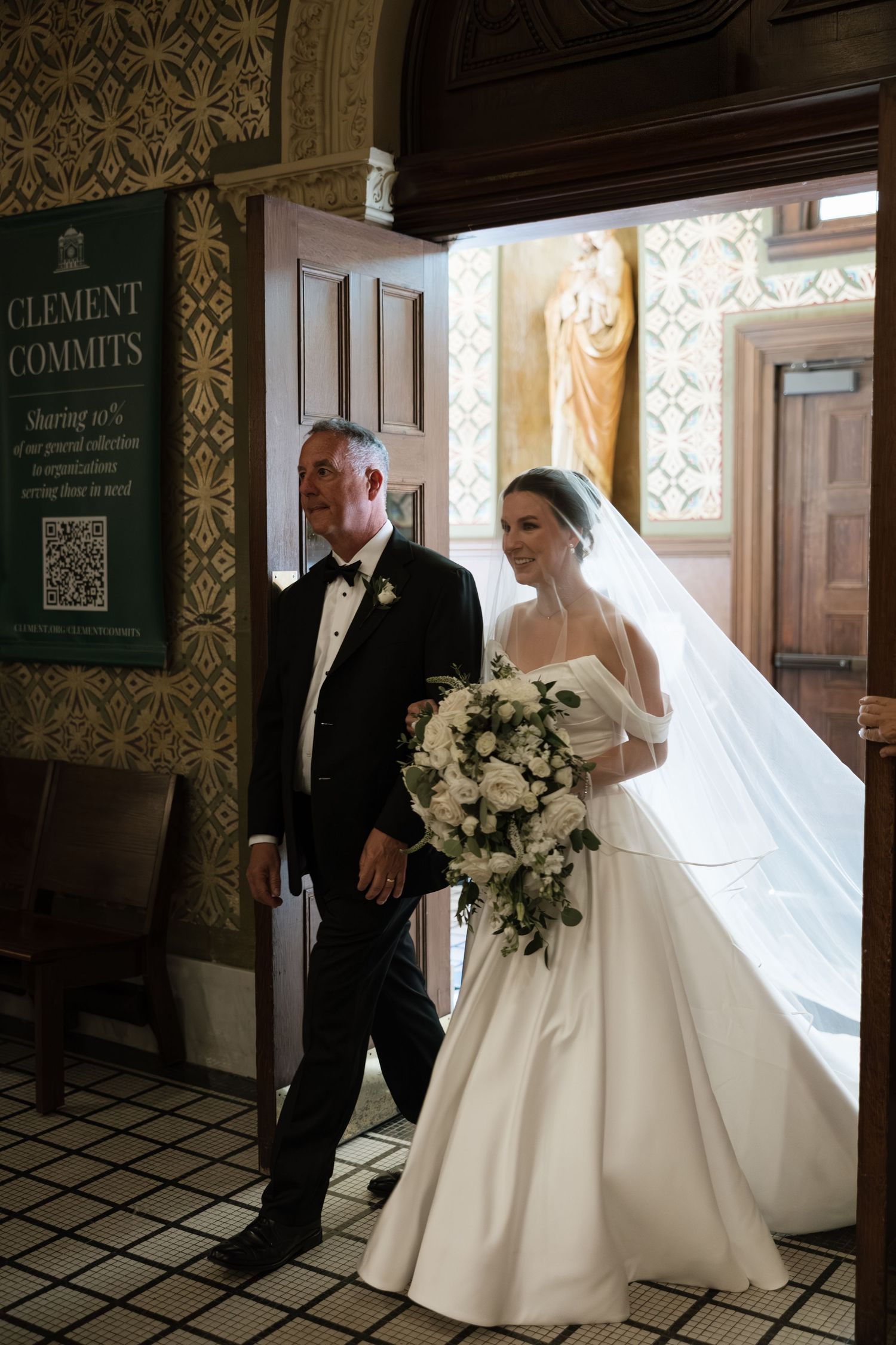 A bride in an elegant white ballgown and veil stands with a suited companion in an ornate hallway with patterned wallpaper.