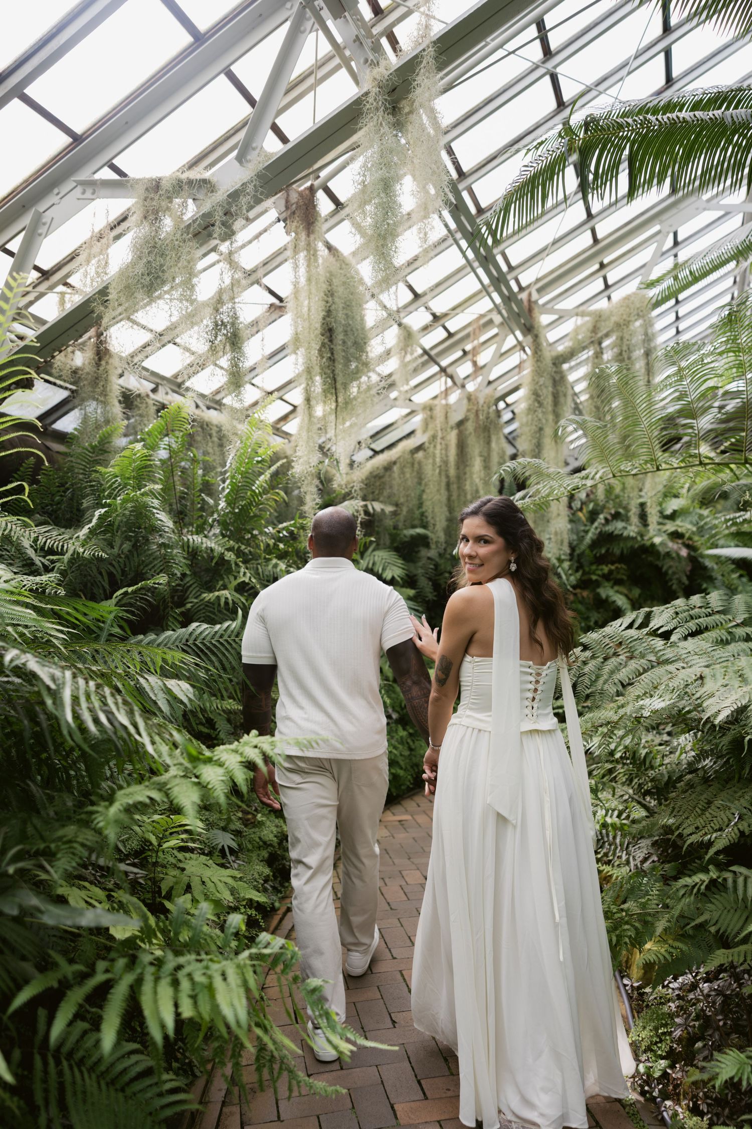 A couple dressed in white walks through a lush greenhouse filled with tropical ferns and palms.