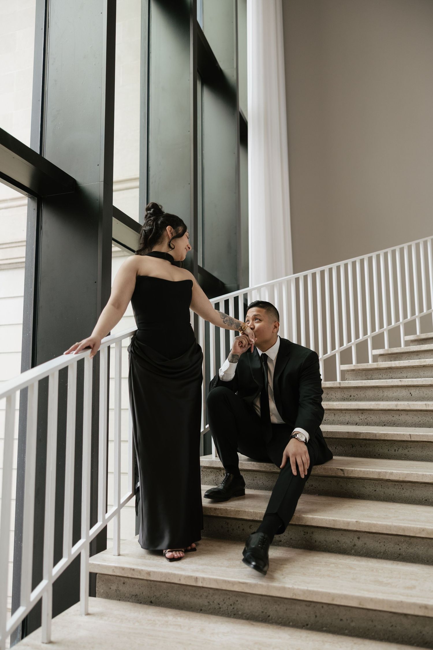 A couple in black attire sits together on modern concrete stairs in an artistic pose.