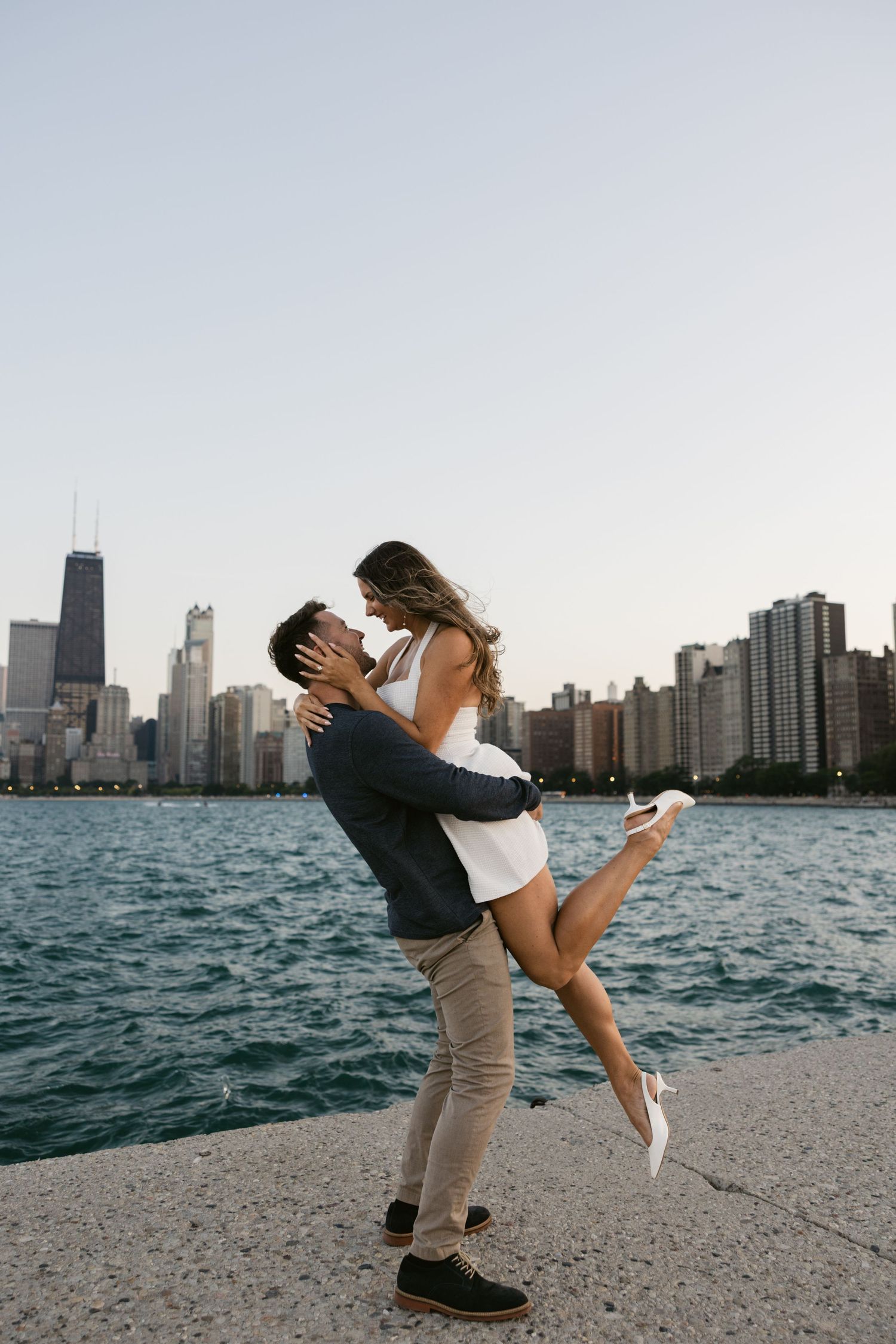 Romantic couple plays and dips on Lake Michigan shoreline with Chicago skyline backdrop at sunset.