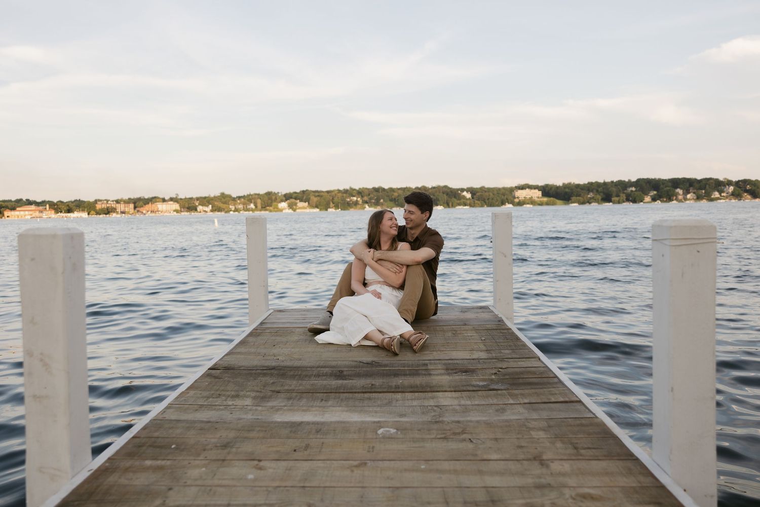 Two people share a romantic moment sitting together on a wooden dock overlooking the water at sunset.