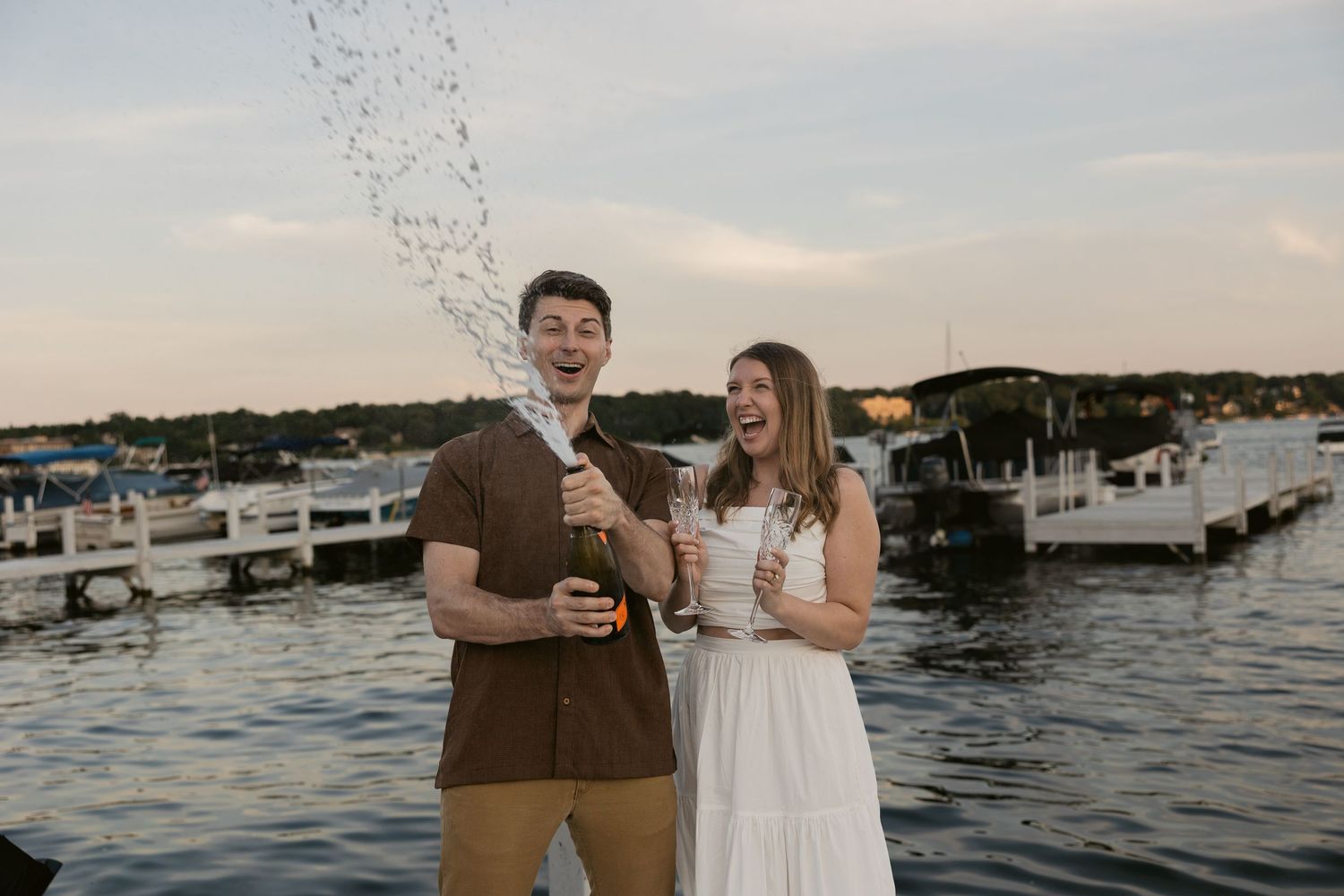 Couple celebrating with champagne on dock with boats in background.