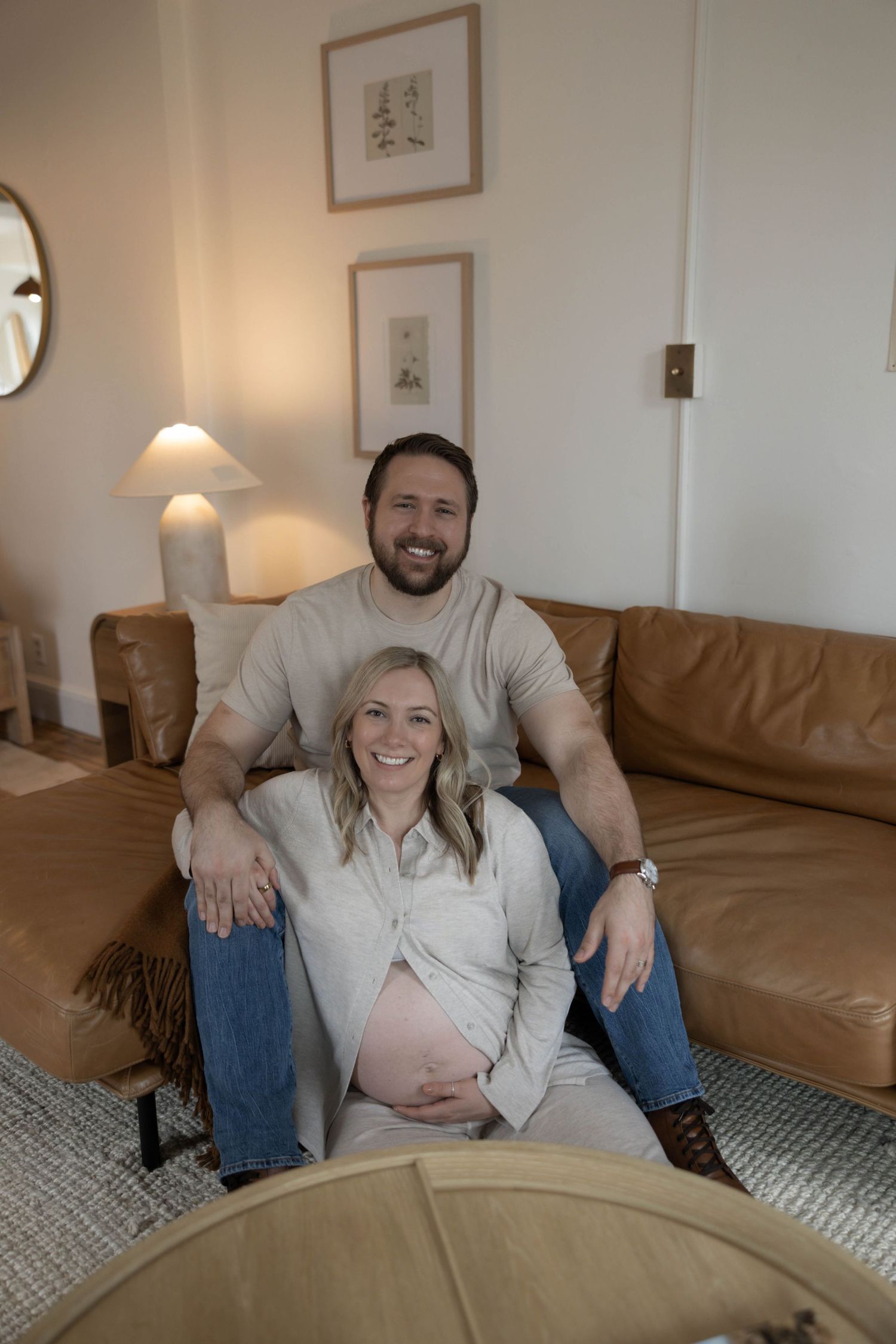 Expectant couple relaxes together on a tan leather couch in a cozy living room with warm lighting.