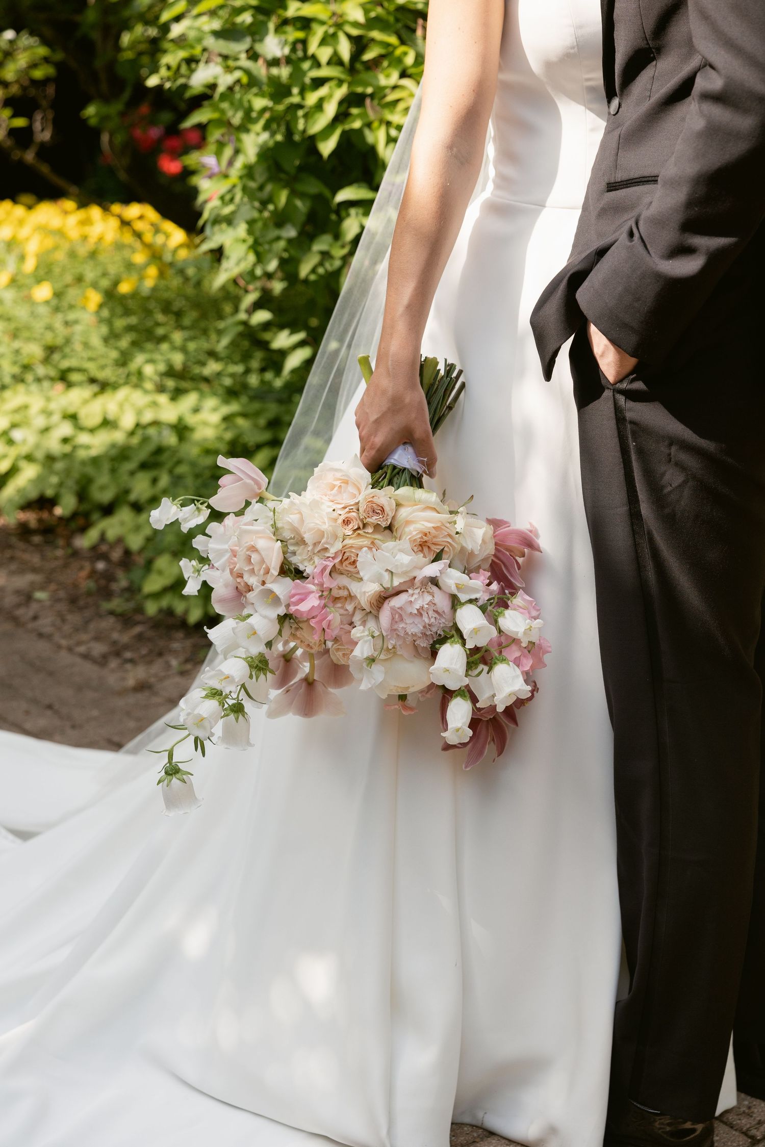 A delicate blush and cream wedding bouquet is held against a white bridal gown and dark suit.