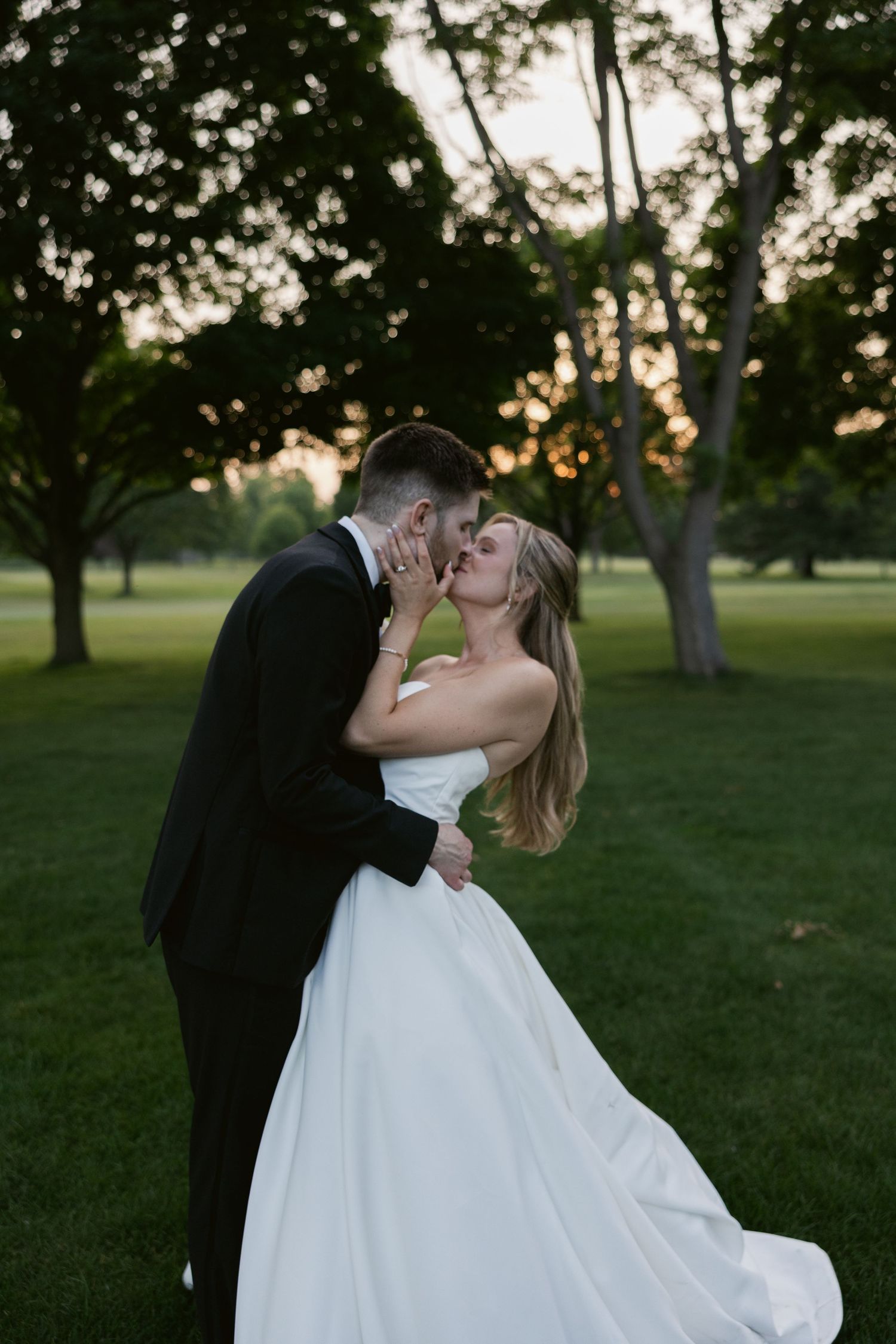 Romantic kiss between newlyweds on grass lawn at sunset.