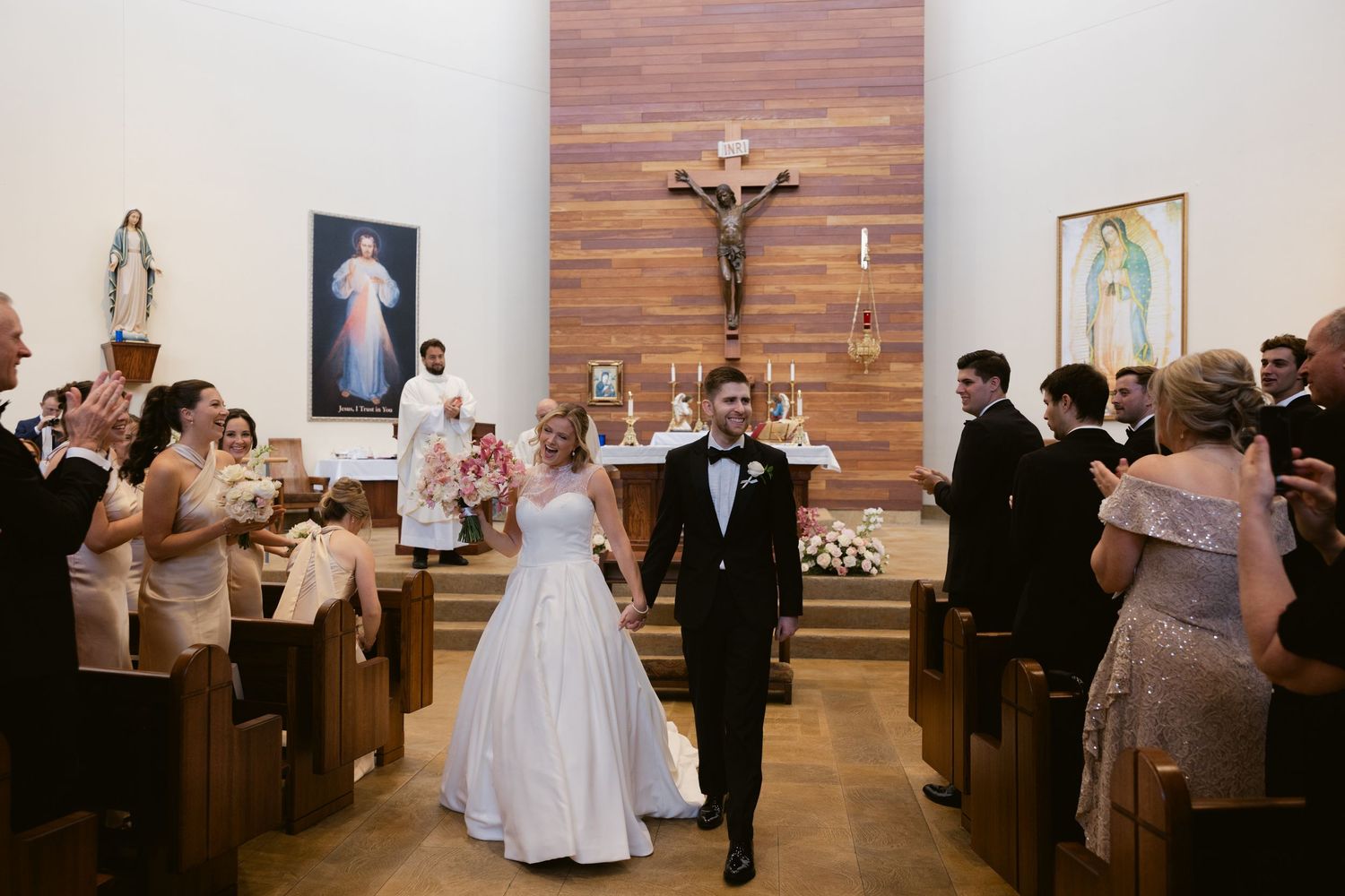 A couple walks down the aisle of a Catholic church after their wedding ceremony while guests celebrate.