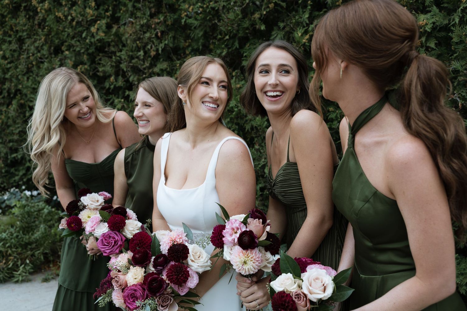 Bridesmaids in dark green dresses hold pink and burgundy bouquets while smiling together outdoors.