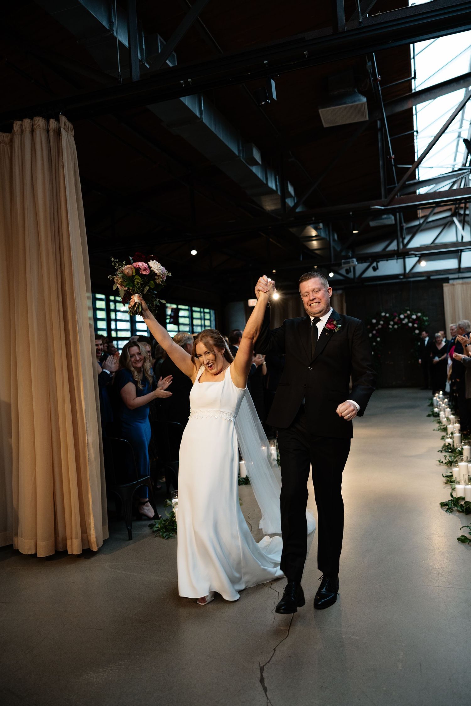 A couple dances joyfully in an industrial-style venue with high ceilings and cream curtains during their wedding celebration.