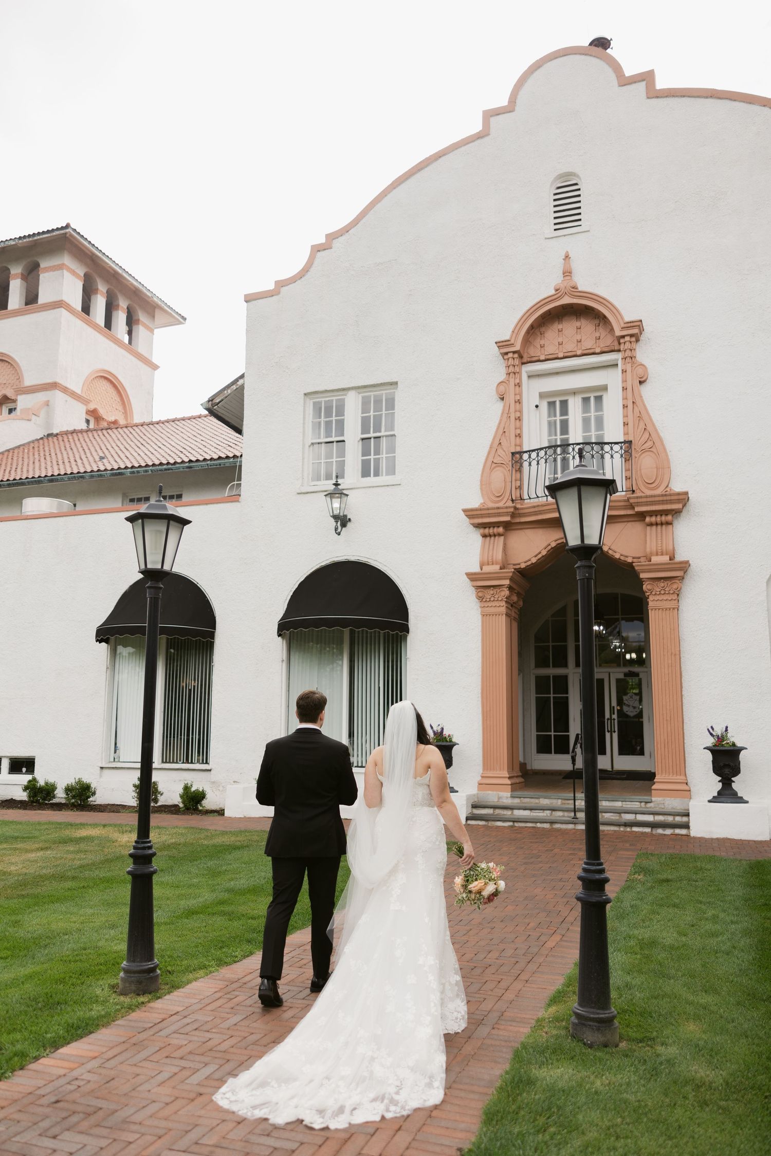 A couple in formal wedding attire walking along a path towards an elegant Spanish colonial style church building.