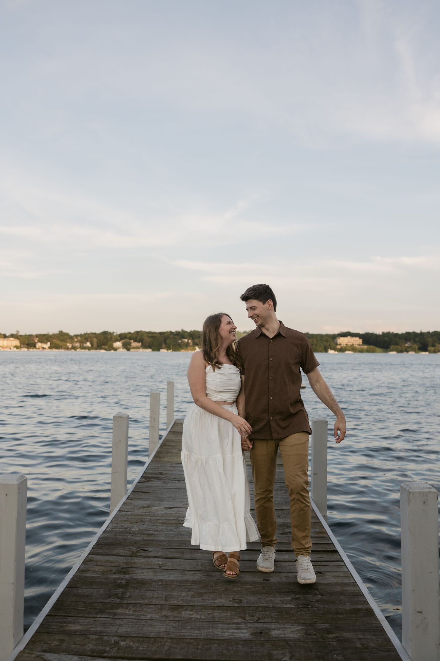 Couple sits together on a wooden dock overlooking a serene lake during sunset with coastal homes visible in background.