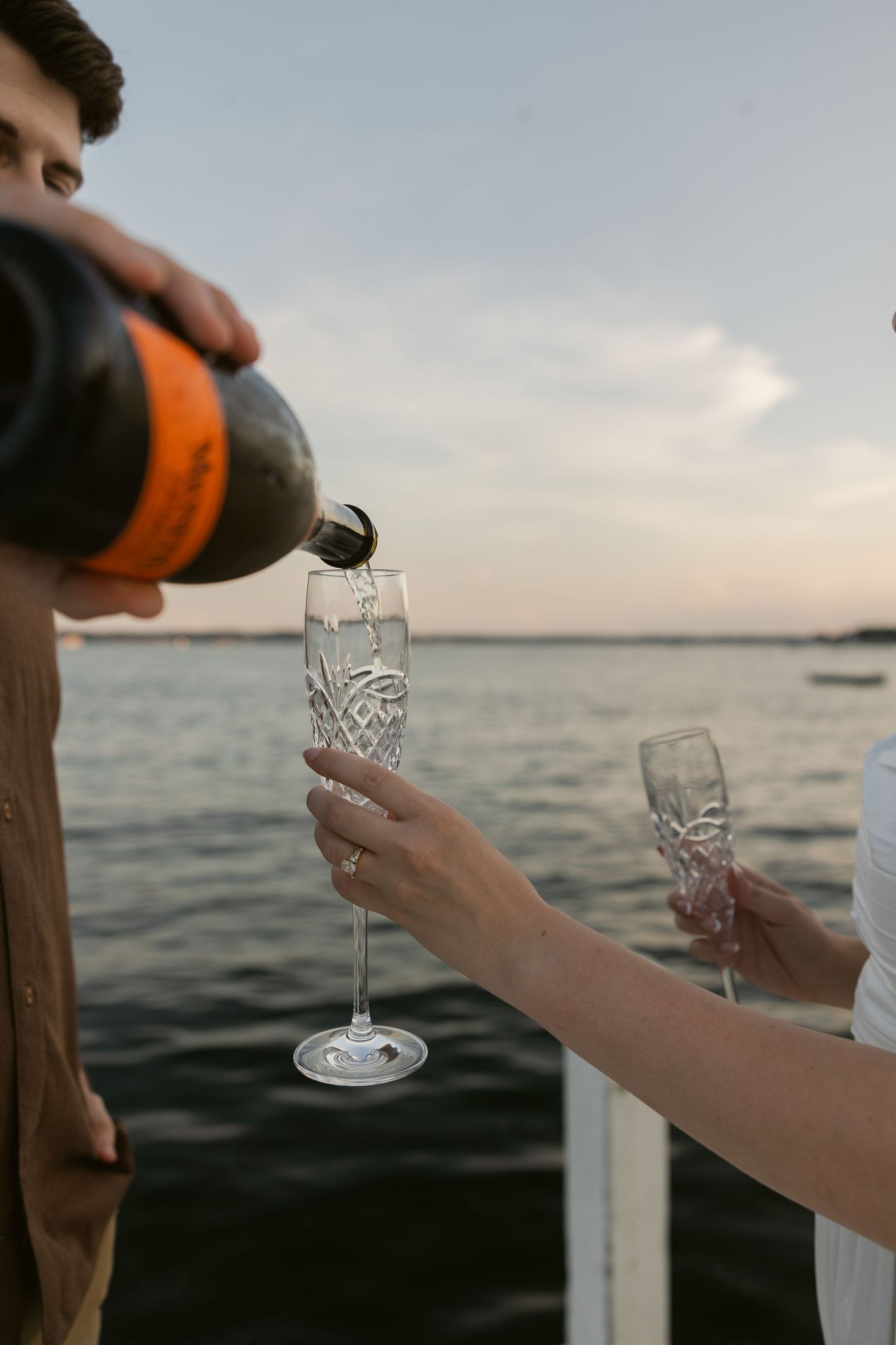 Wine glasses clink together during a sunset toast on a dock by the water.