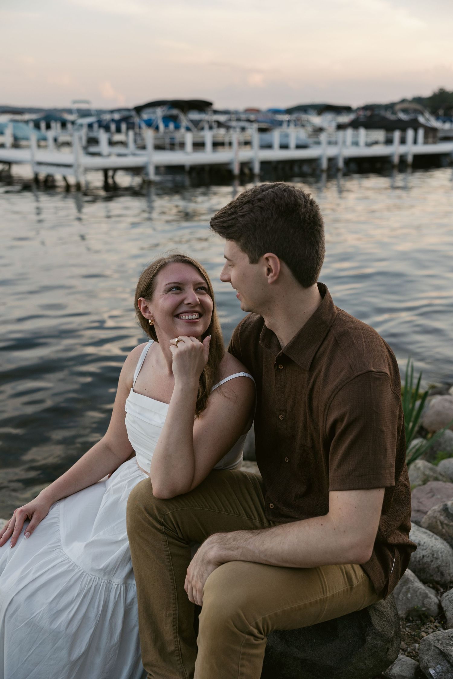 Couple shares intimate moments on a lakeside dock during golden hour with boats in background.