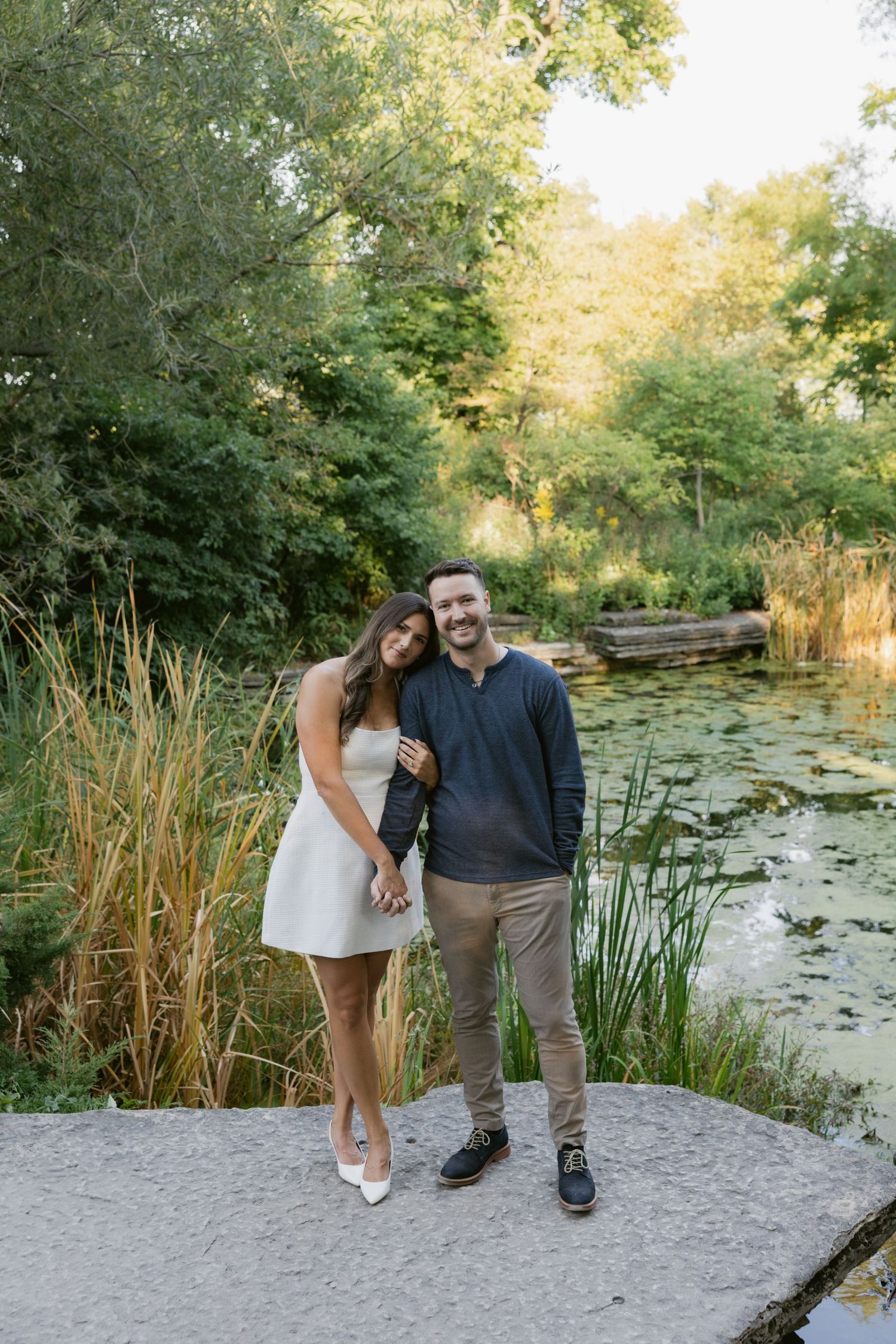 A romantic couple stands near a peaceful pond with lily pads and reeds at sunset.