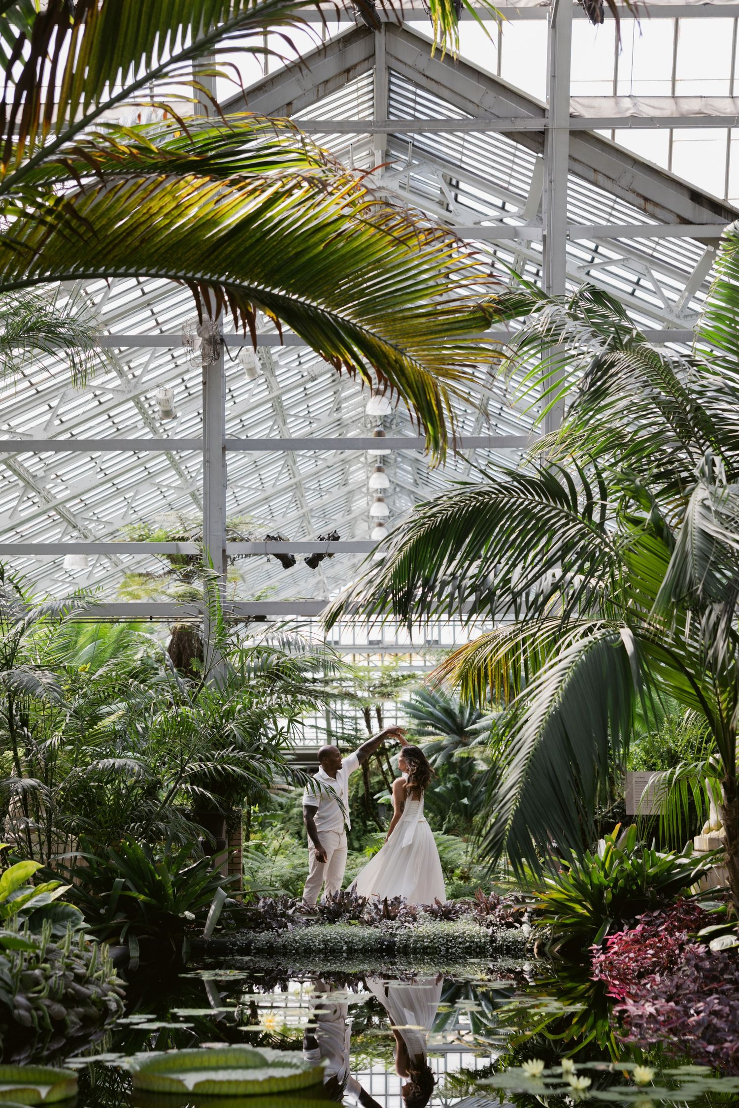 Two people stand together in a lush greenhouse conservatory with tropical palms and a reflecting pool.