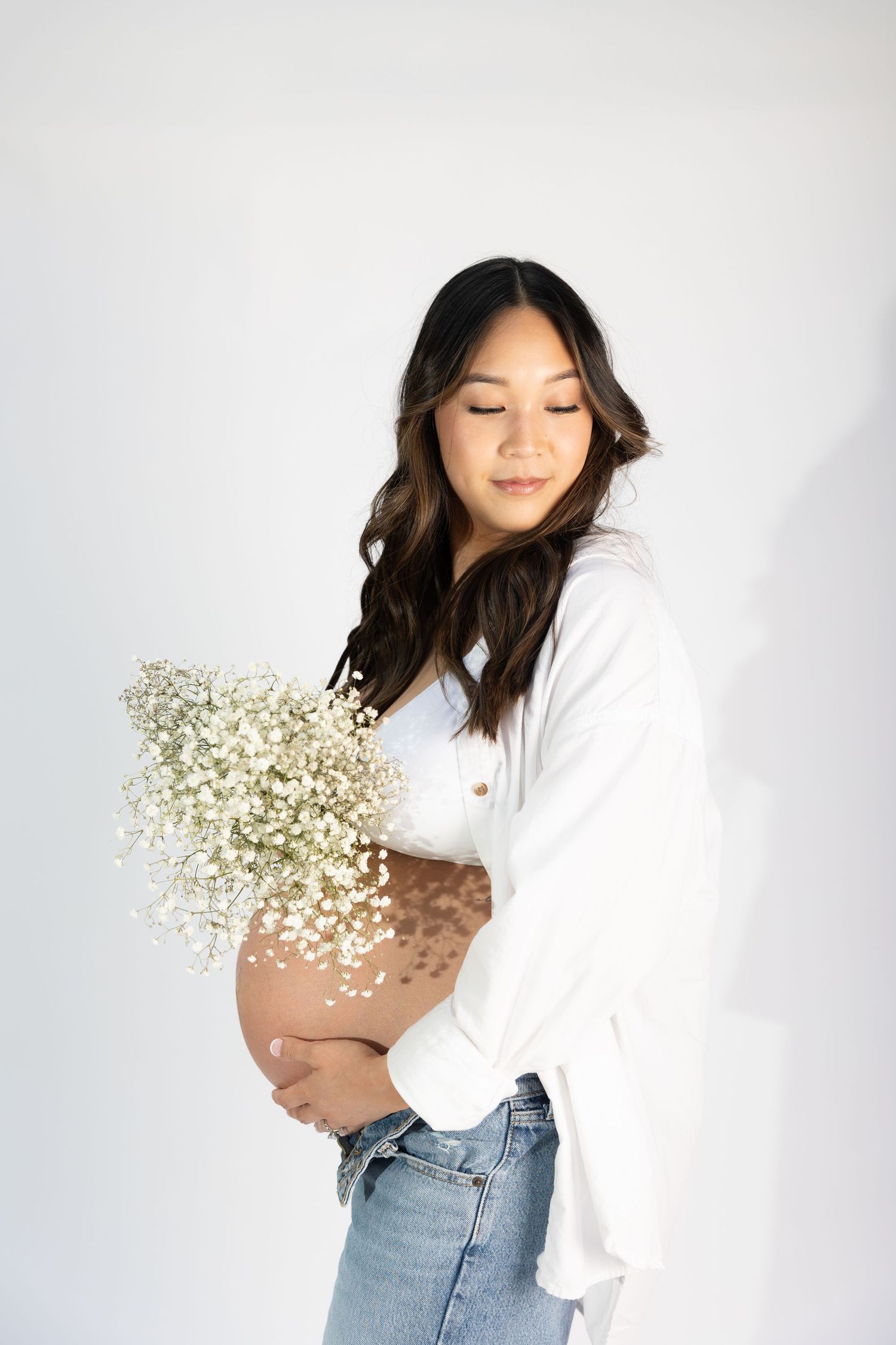 Serene maternity portrait with woman in white holding flowers against light background.