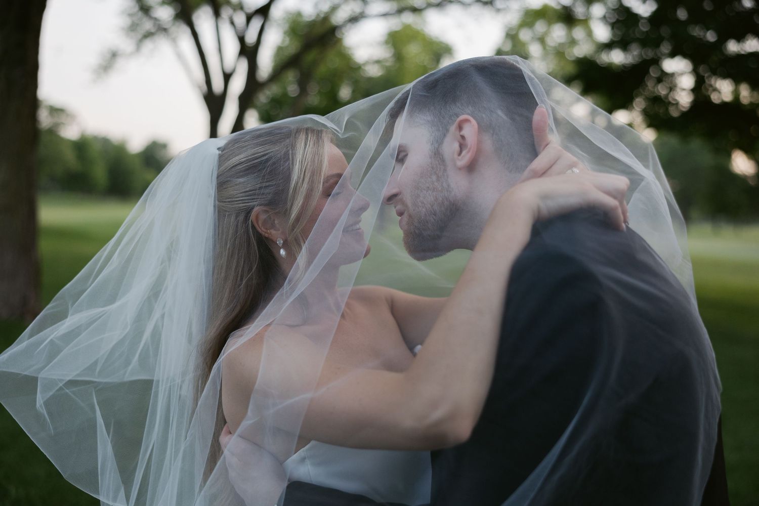 A romantic wedding couple shares an intimate moment beneath a flowing veil in an outdoor garden setting.