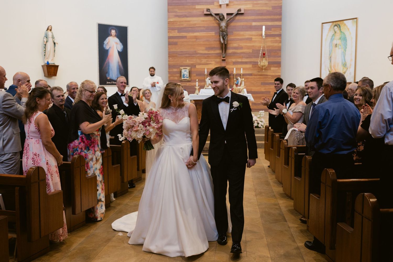 Newlyweds walk down the wooden aisle of a church while guests cheer from the pews.