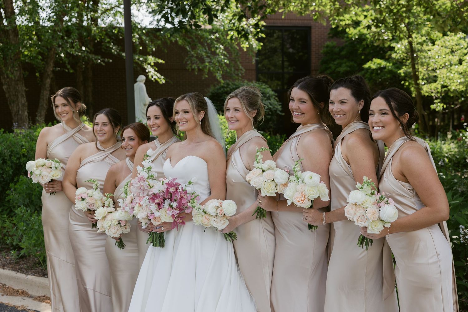 A bridal party in champagne dresses poses with bouquets in a garden setting.