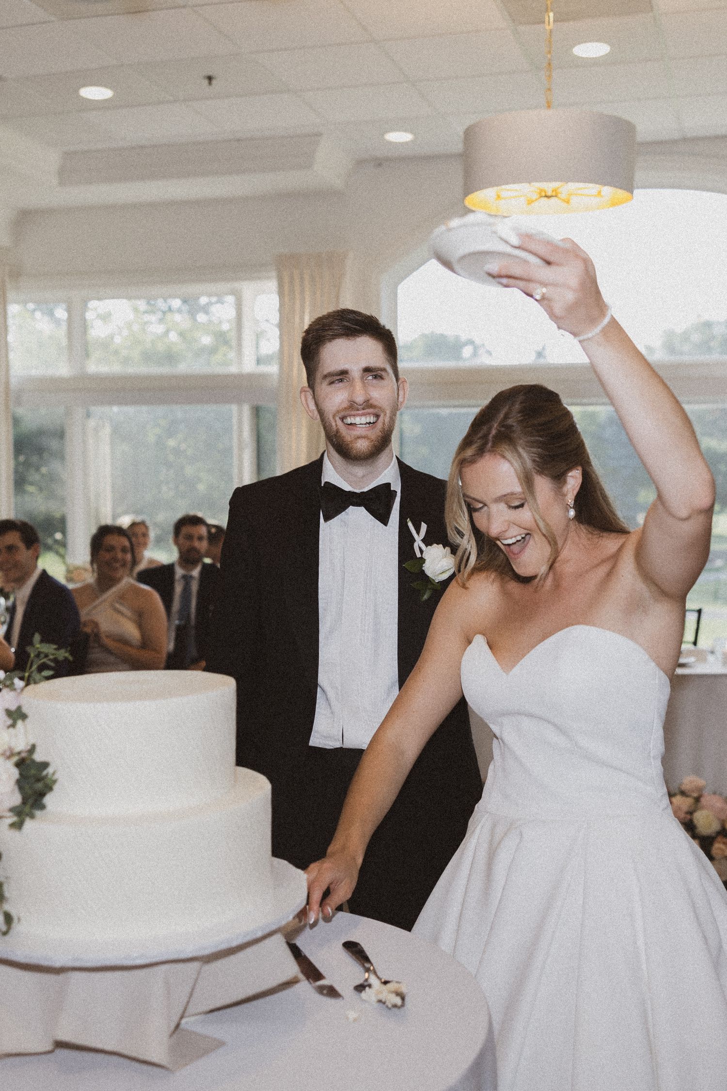 Newlywed couple cuts their elegant white wedding cake during reception as bride laughs joyfully.