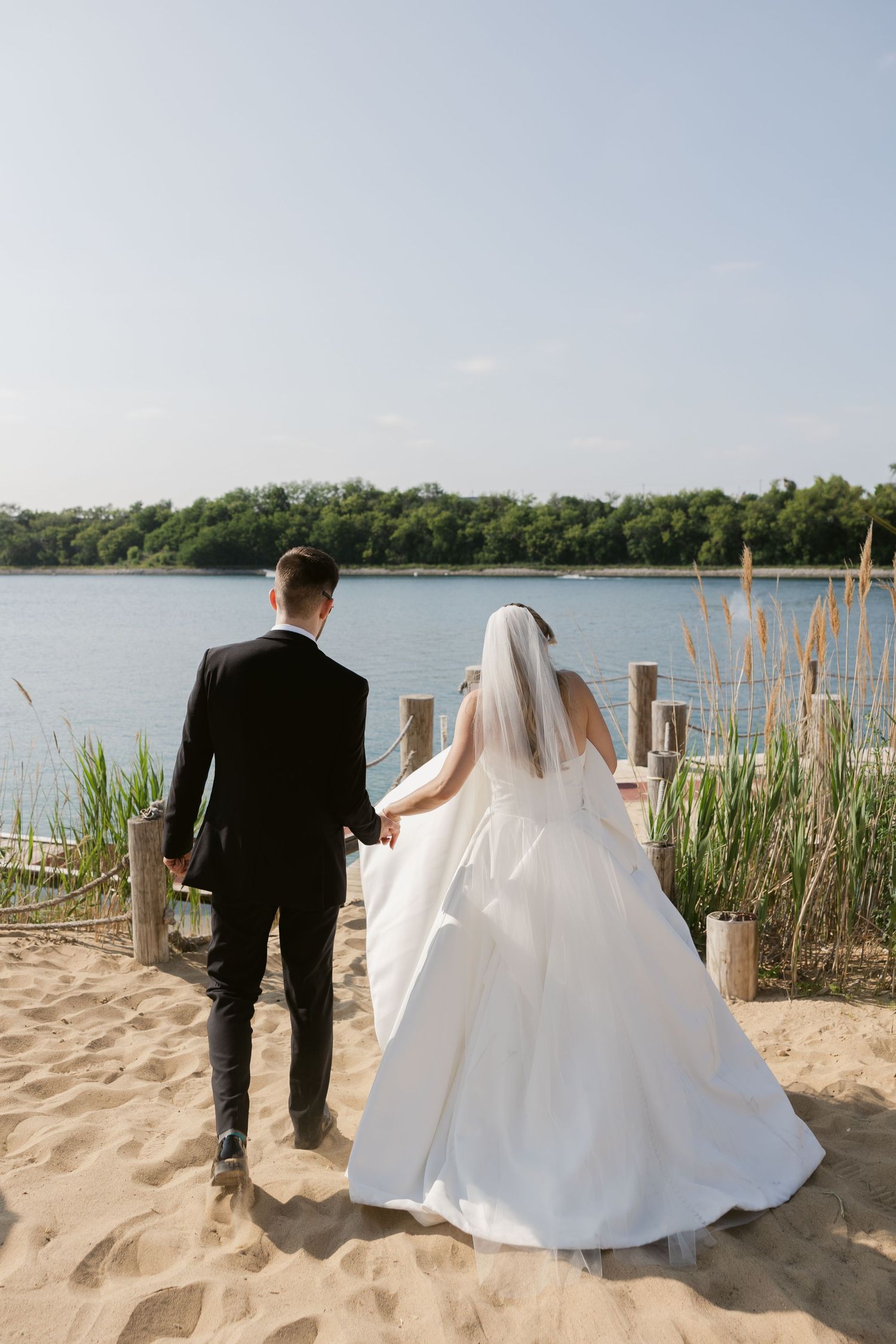 Newlyweds walk along a sandy beach path near cattails and water.
