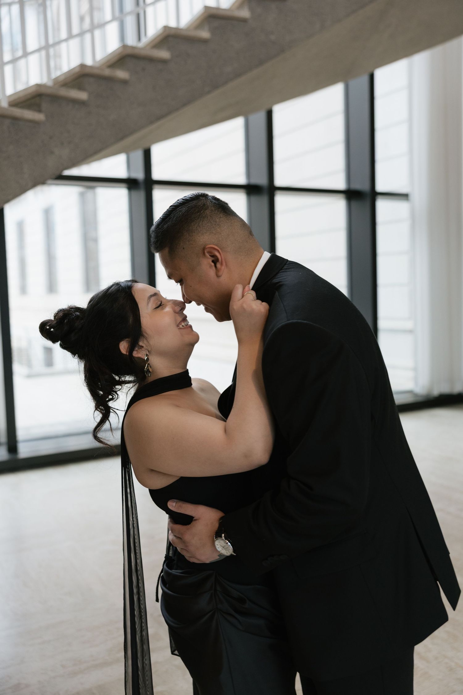 A romantic couple in formal black attire share an intimate dance pose in a bright modern interior space.