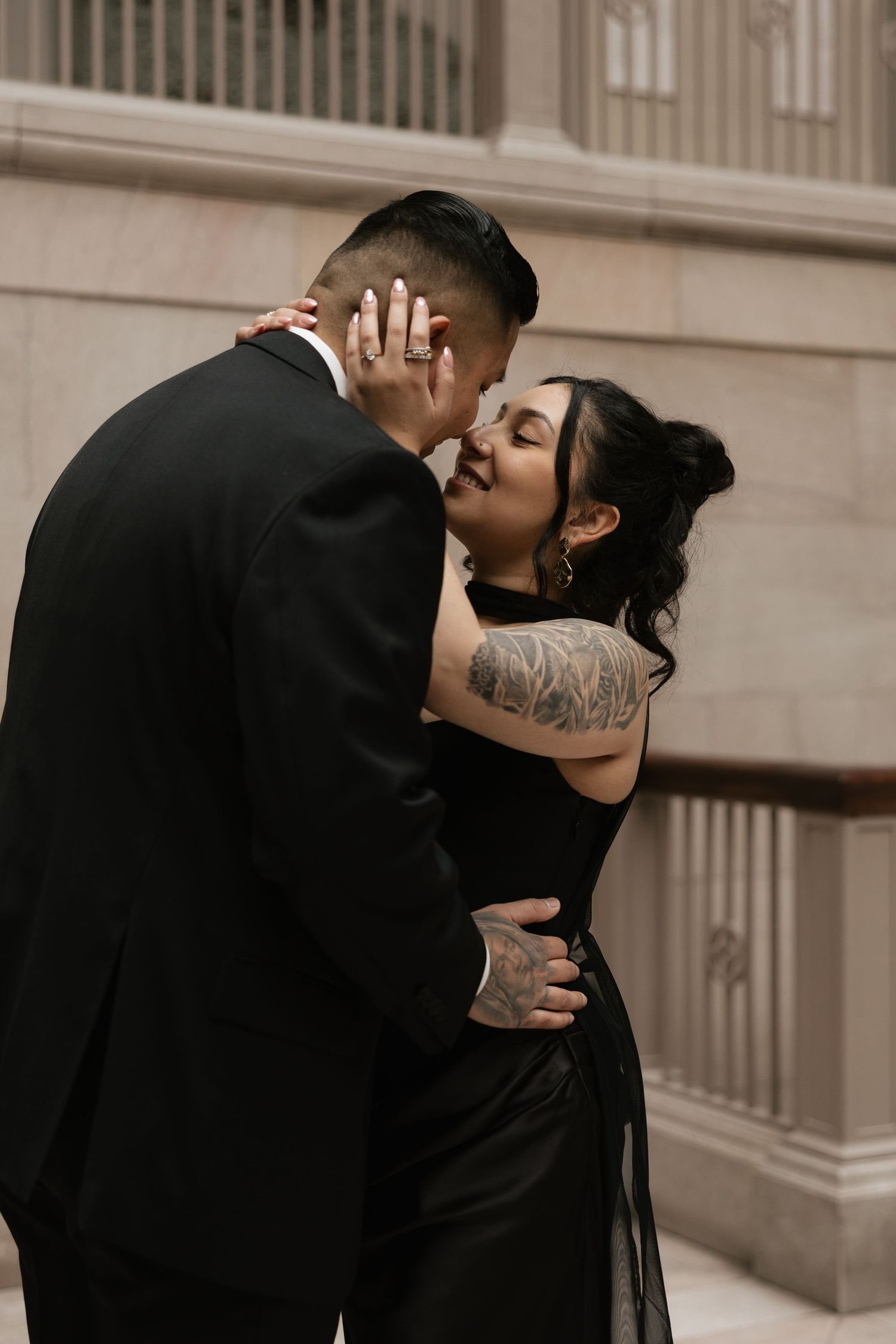 A romantic couple shares an intimate moment against a beige wall in formal attire.