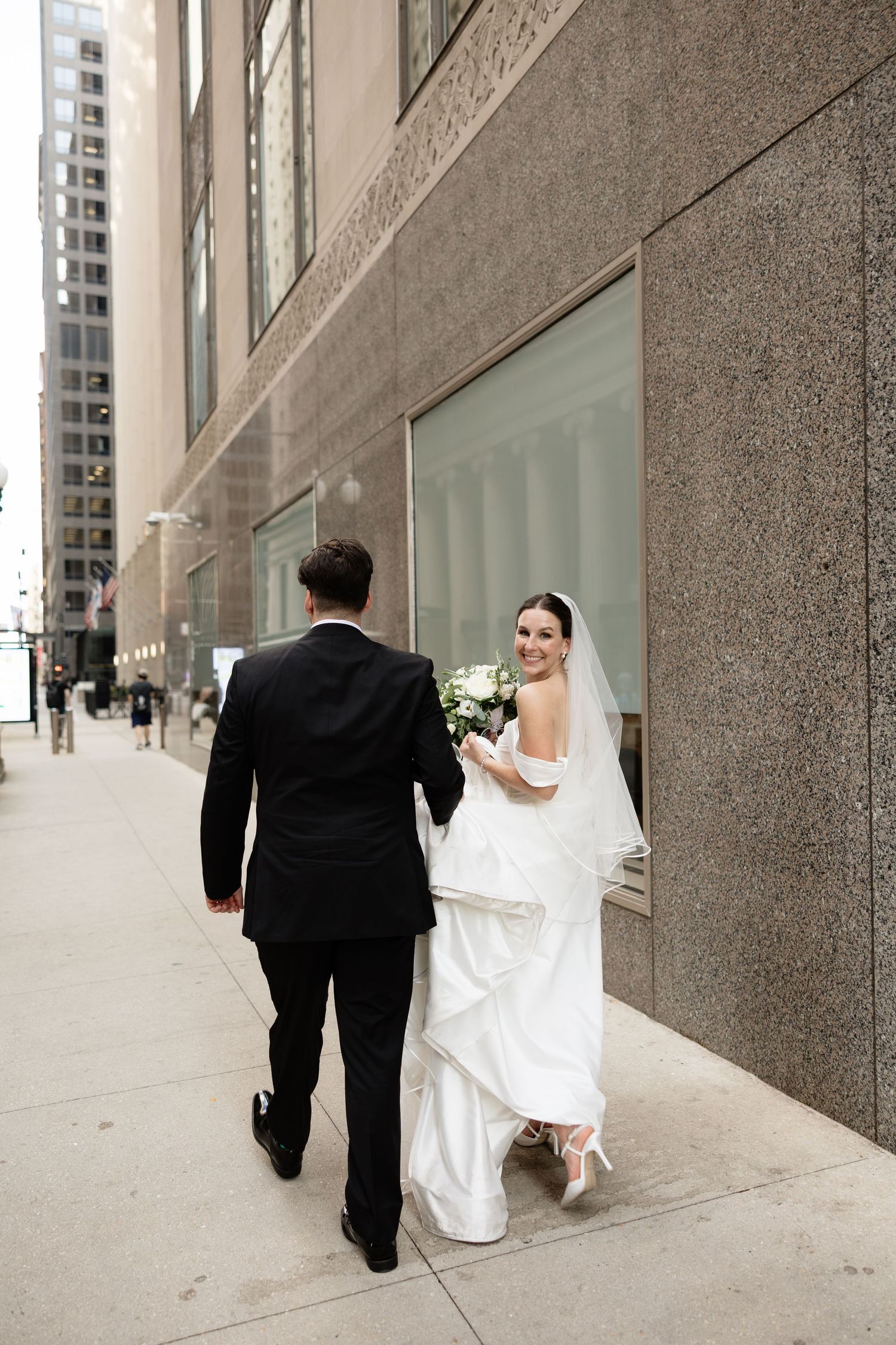 A bride in white and groom in black walk down a modern urban sidewalk with concrete walls.
