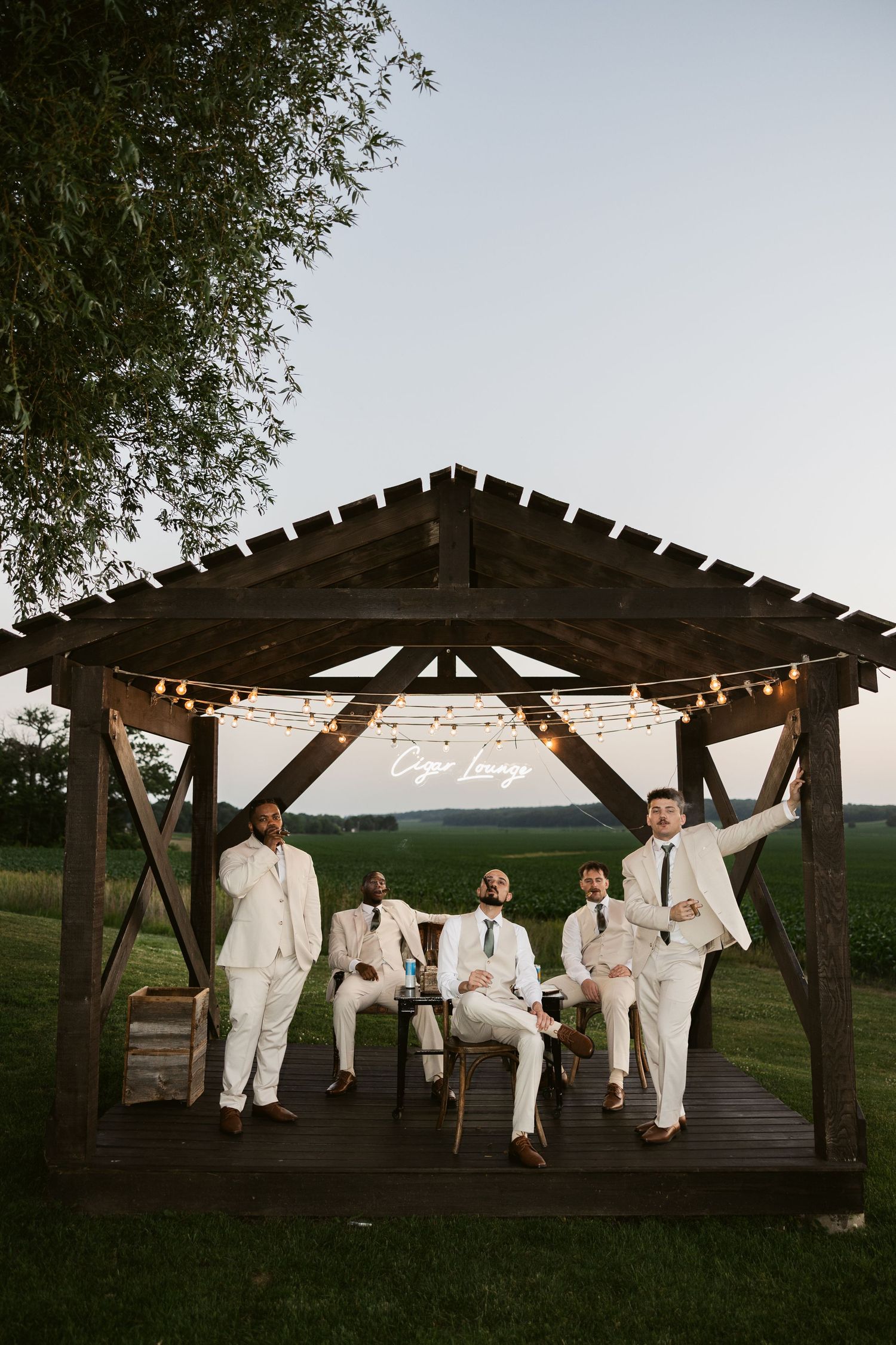 A group of people in crisp white suits pose under a wooden gazebo decorated with string lights at sunset.