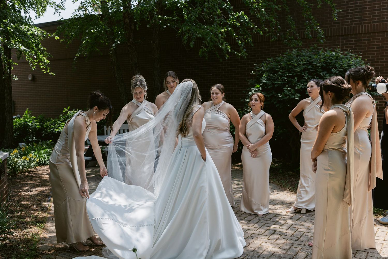 Wedding party in neutral dresses gathered outdoors on brick pathway under green foliage.
