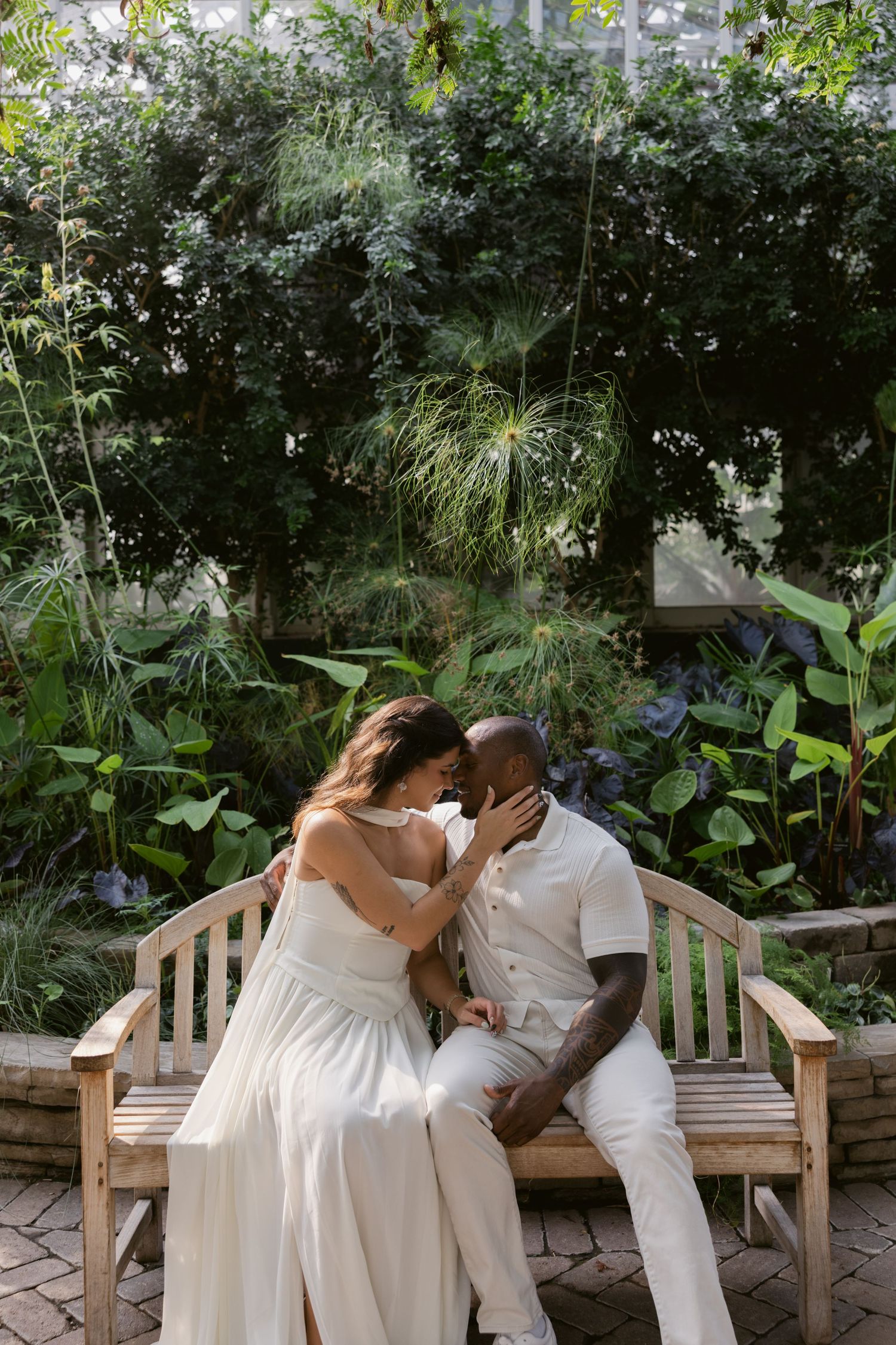 A couple shares a tender moment on a wooden bench surrounded by lush garden greenery.