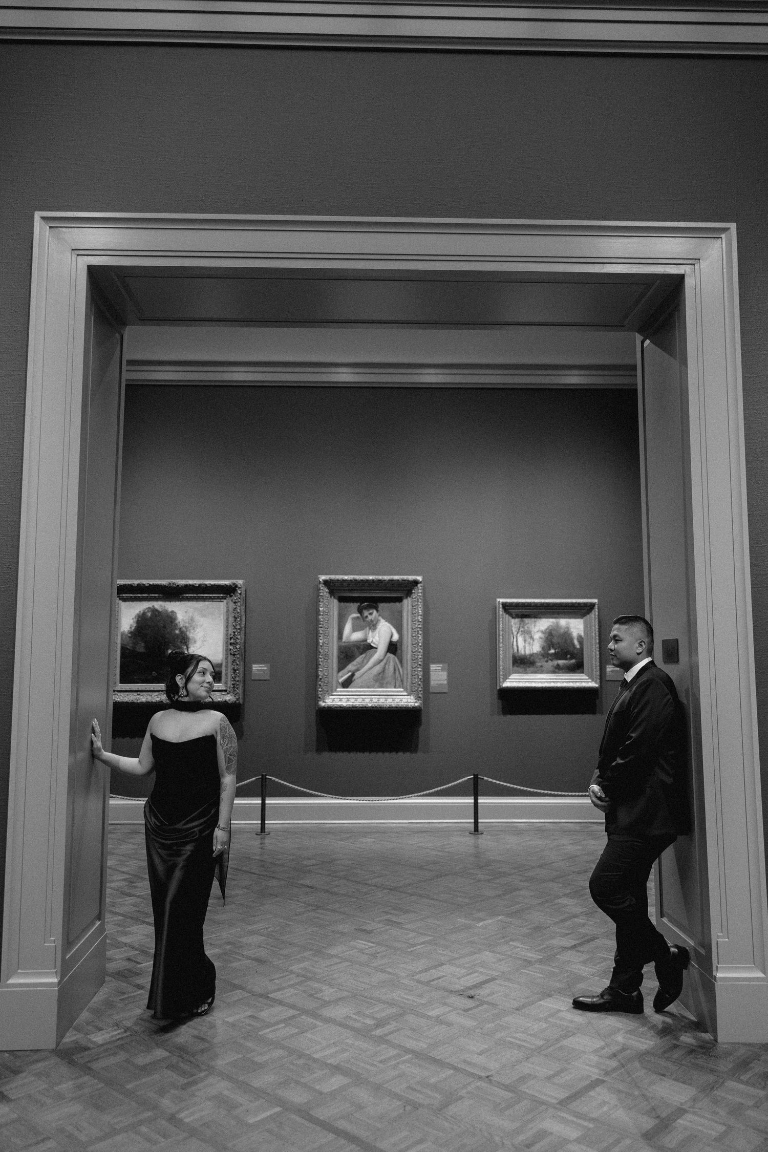 A couple explores a museum gallery with framed artwork and parquet floors in black and white.
