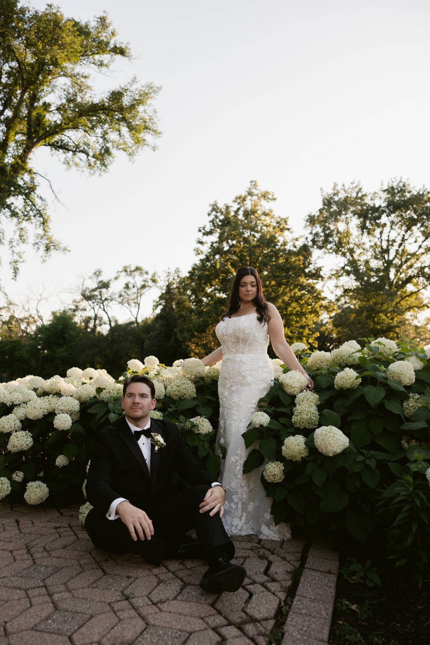 A romantic wedding photoshoot in a garden with white hydrangea bushes and stone paving at sunset.