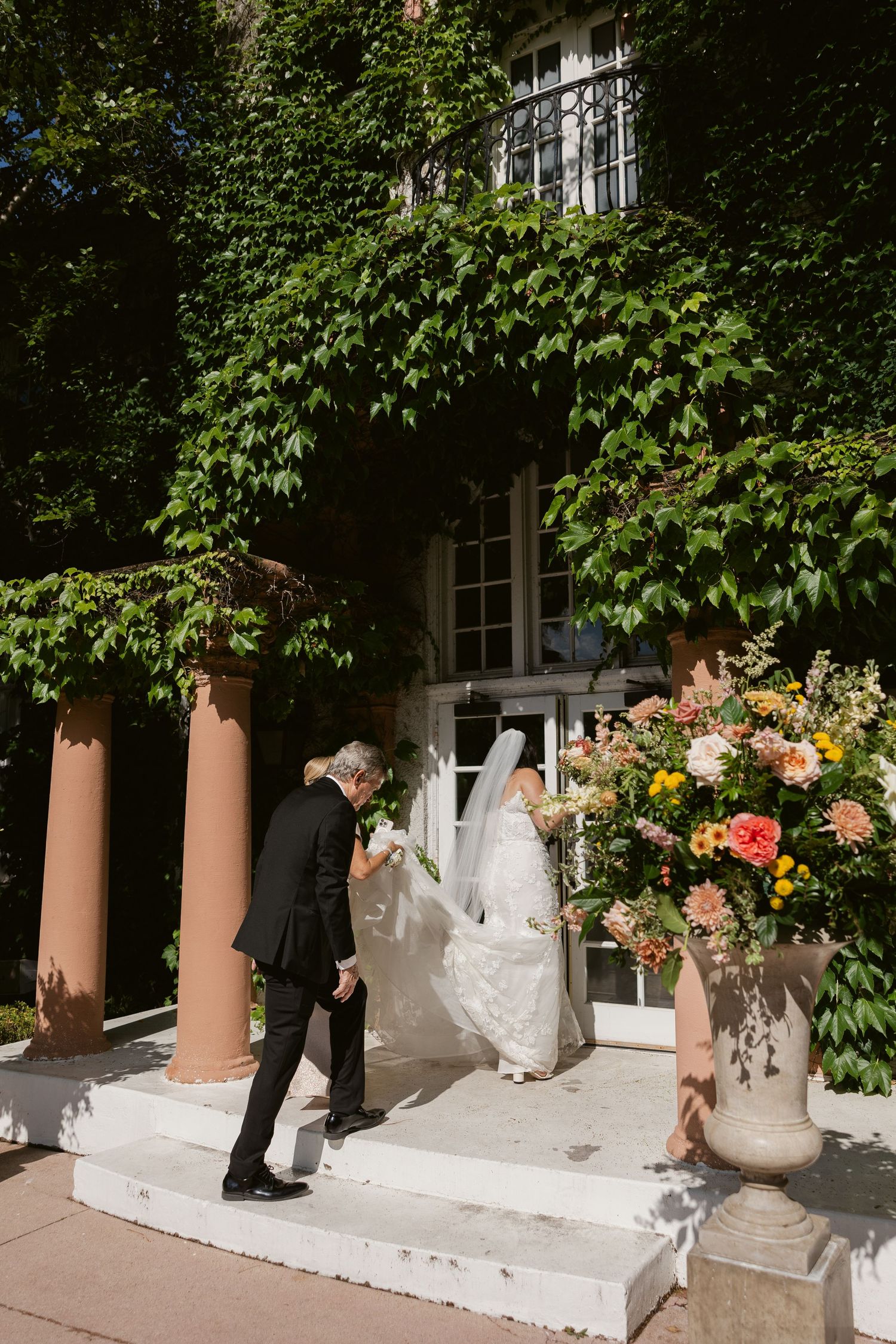 A couple in formal wedding attire ascends stone steps at an elegant ivy-covered venue with floral arrangements.