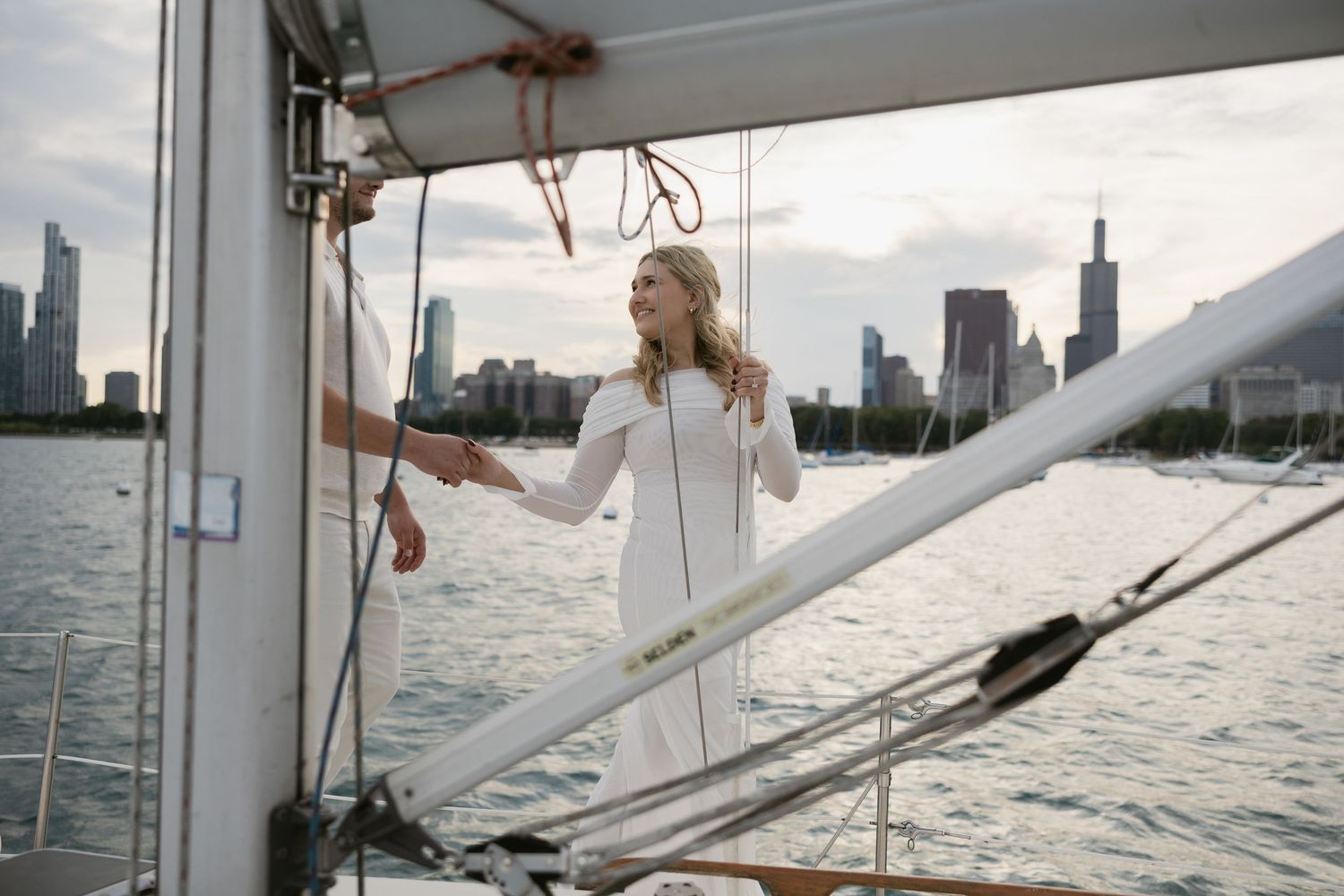 A woman in a white dress explores the deck of a sailboat with the city skyline behind her.