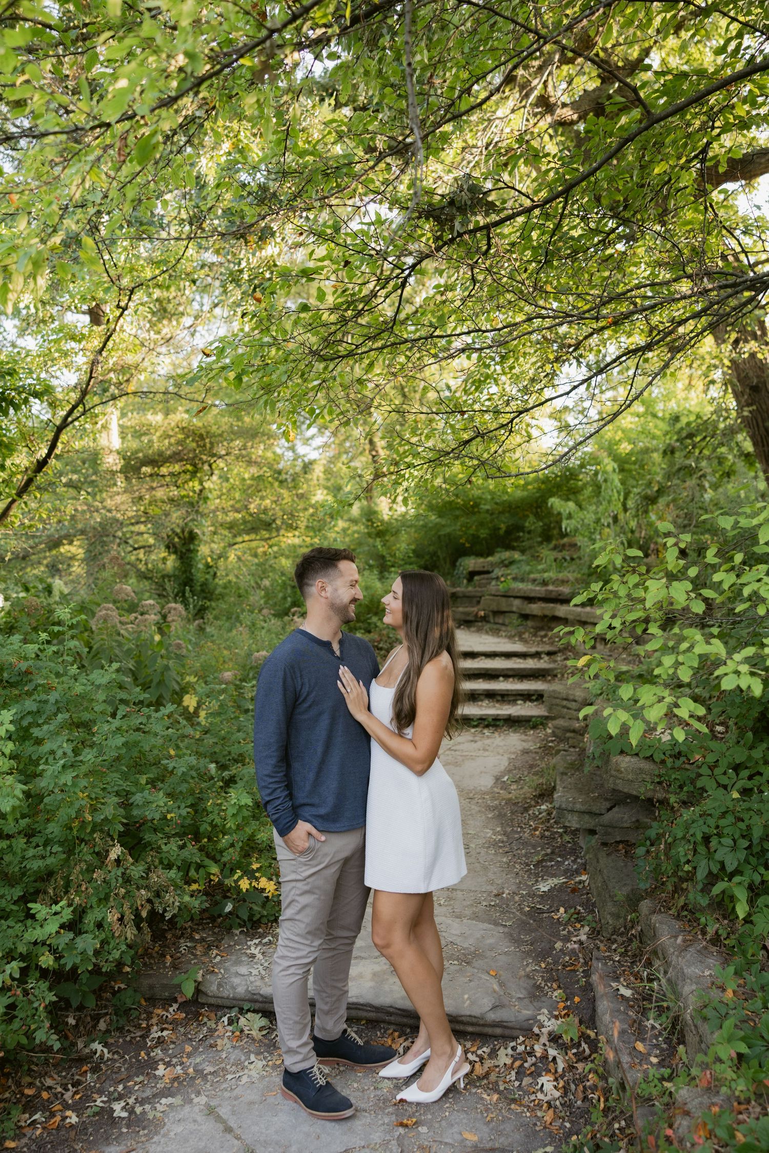 A couple shares a romantic moment on a tree-lined path in a lush green park setting.