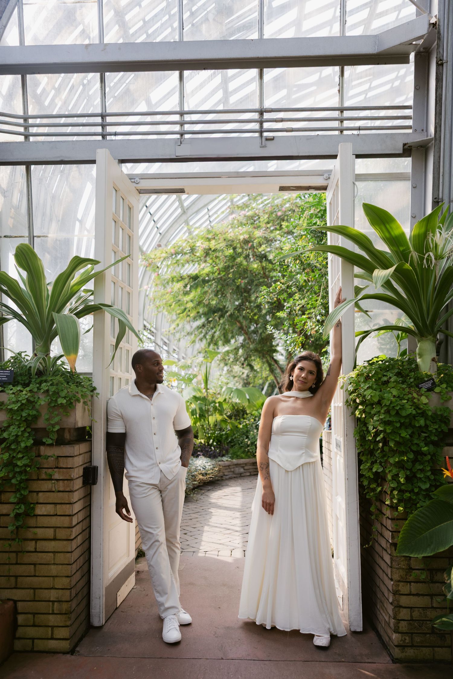 Wedding couple walks through a greenhouse hallway with lush green plants and white walls in a four-panel sequence.