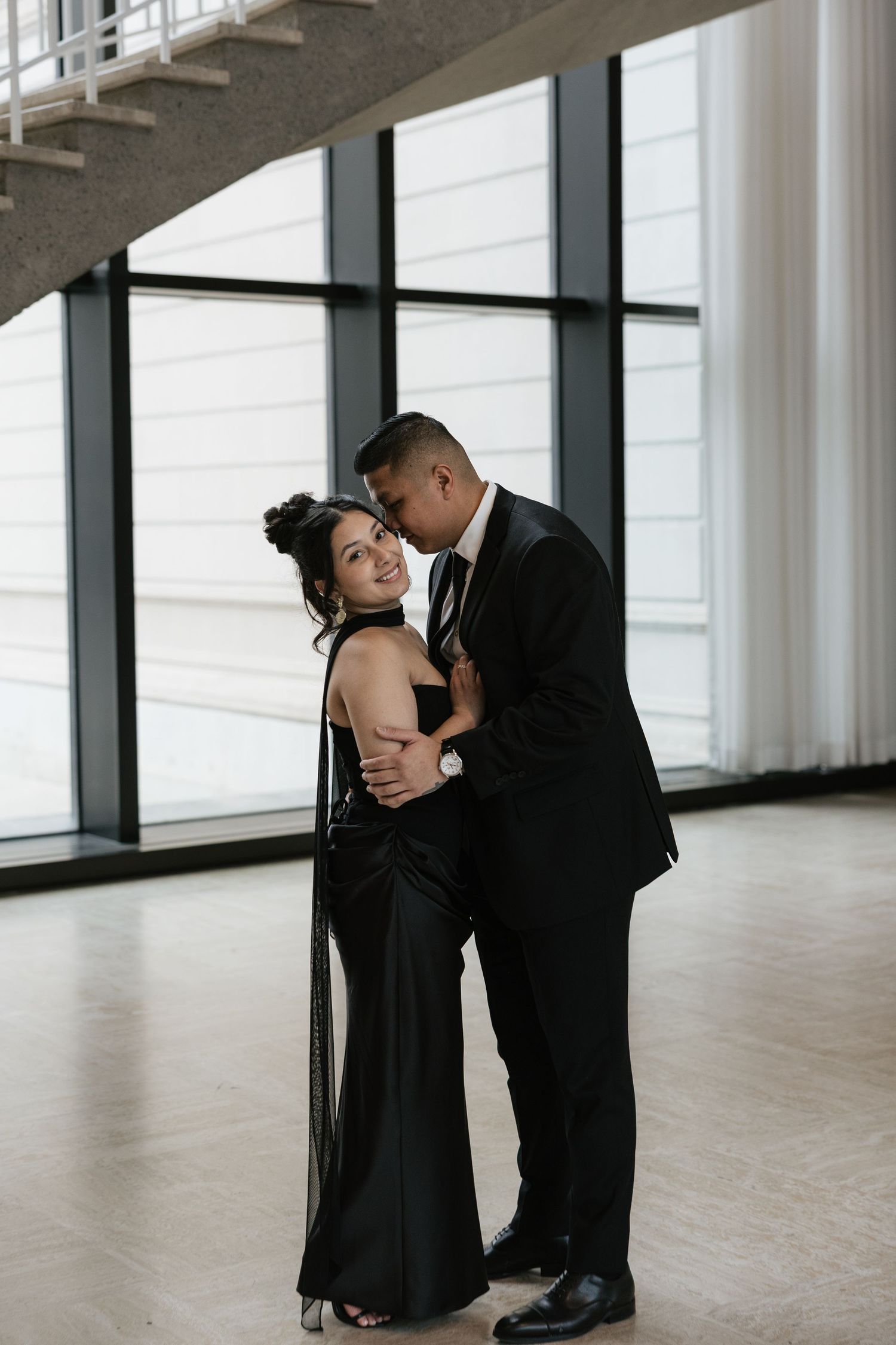 Romantic couple in formal attire embracing in a modern minimalist space with large windows.