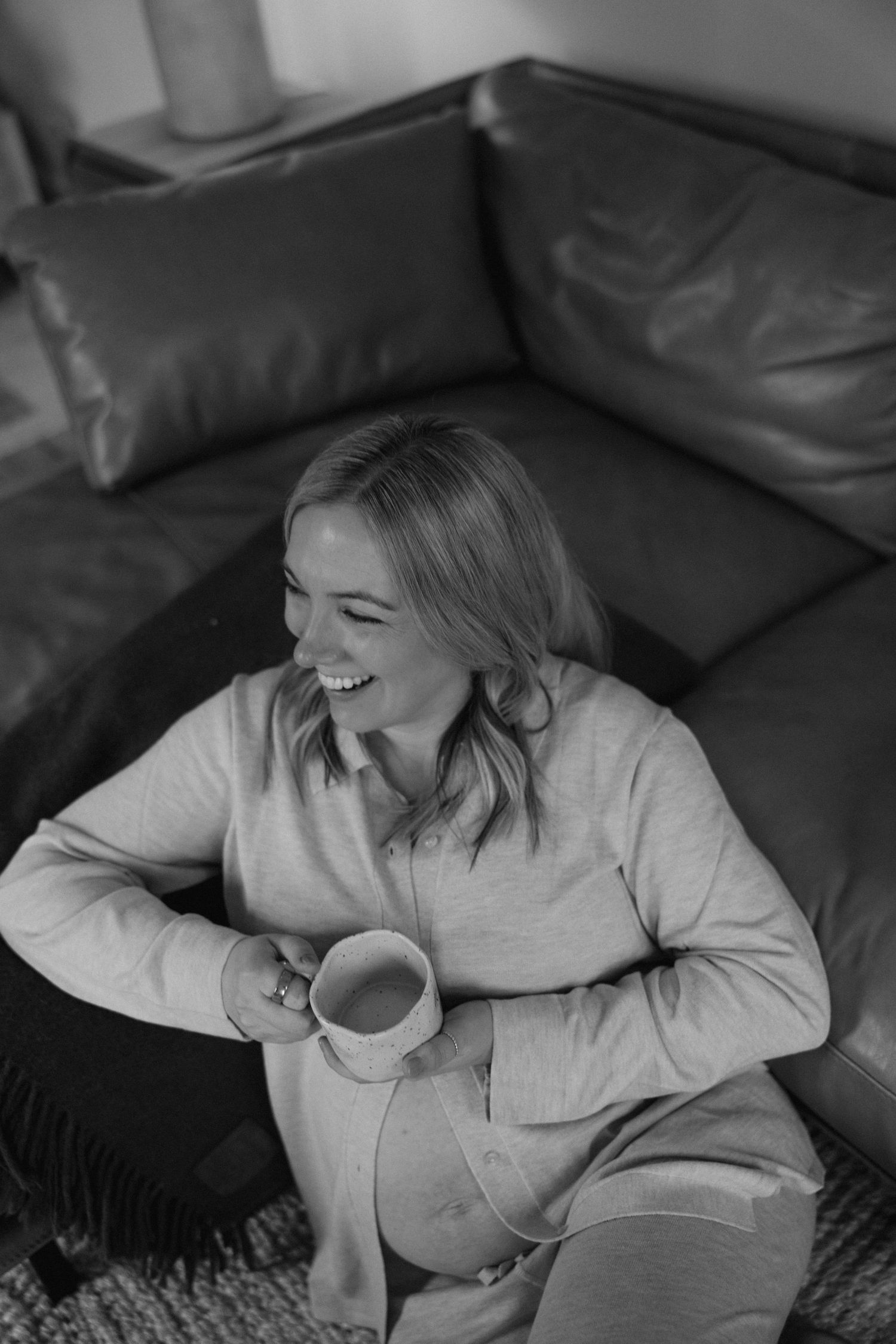 A candid black and white photo shows a joyful moment of relaxation on a leather couch with a warm beverage.