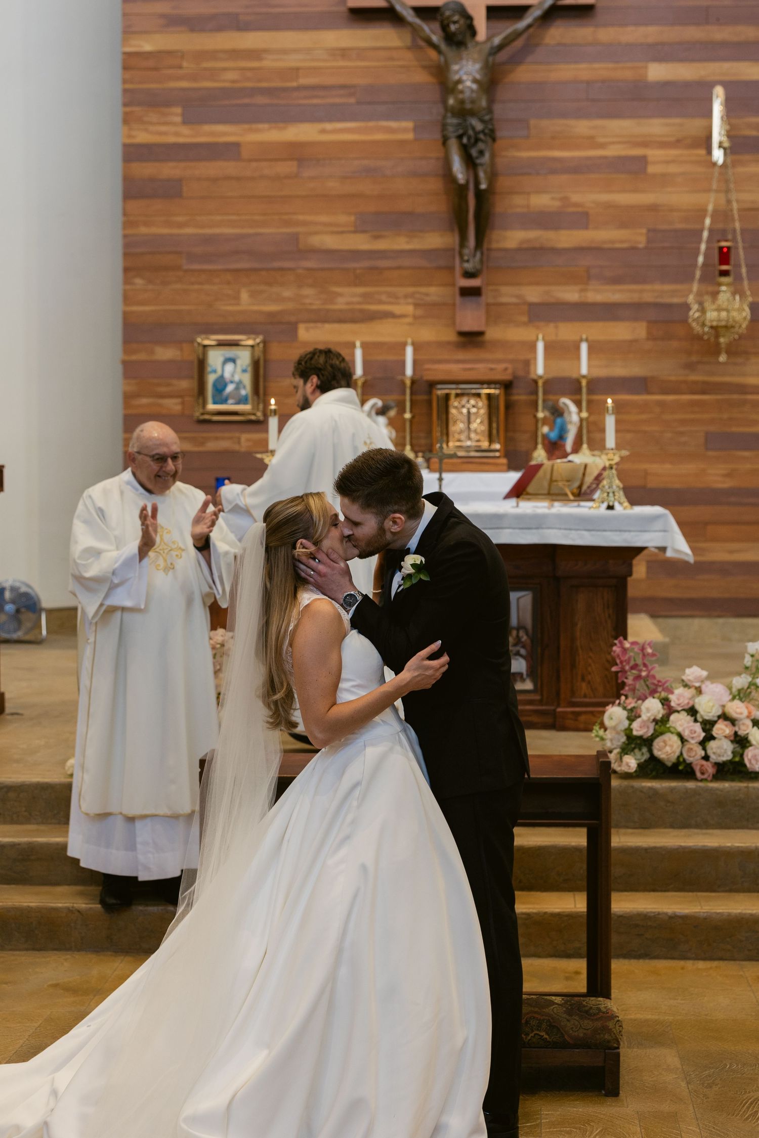 Newlyweds share their first kiss during Catholic church ceremony with priest and wooden cross on wall.