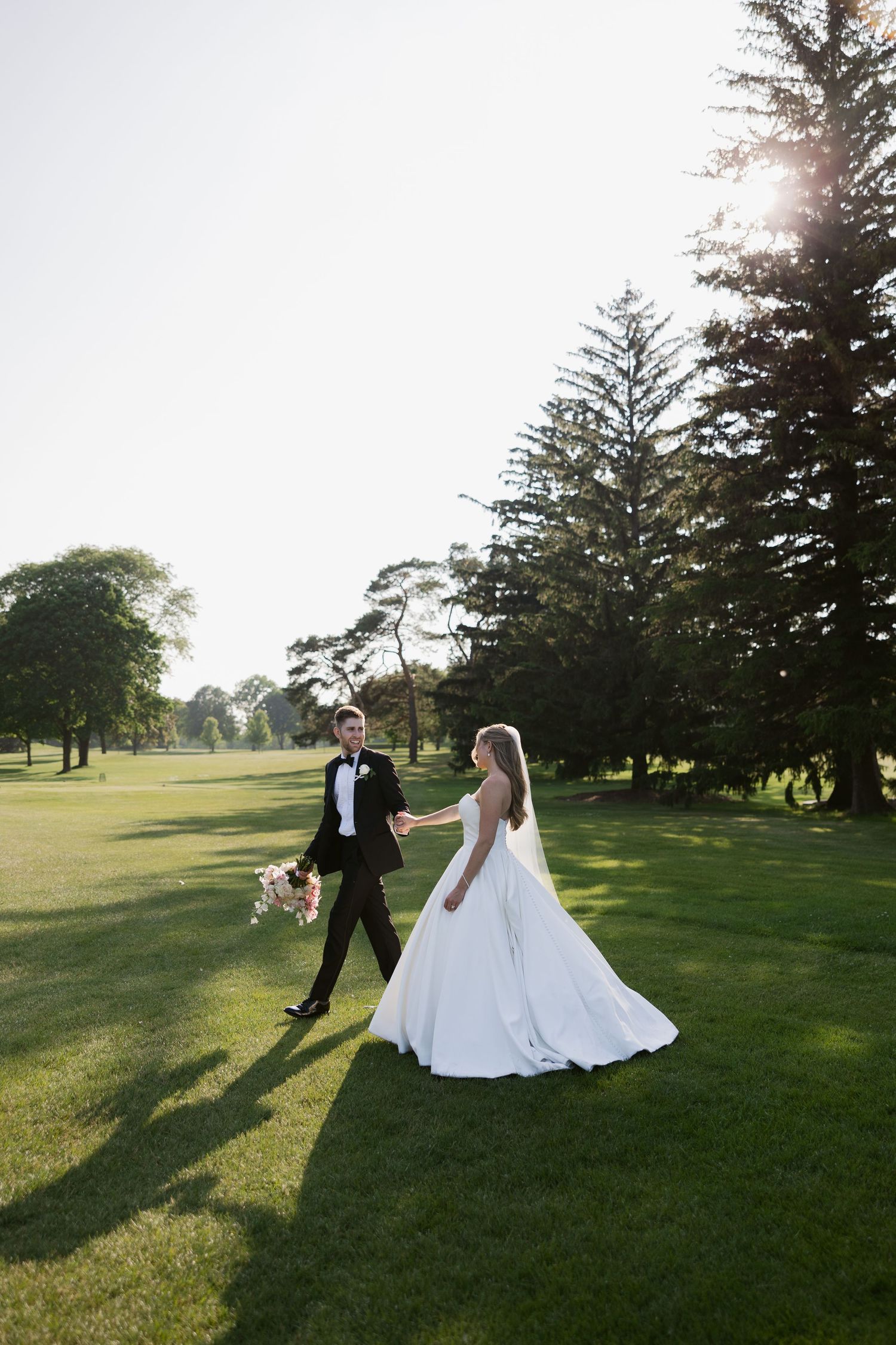 A couple in wedding attire walking and holding hands on a sunlit golf course with evergreen trees in the background.