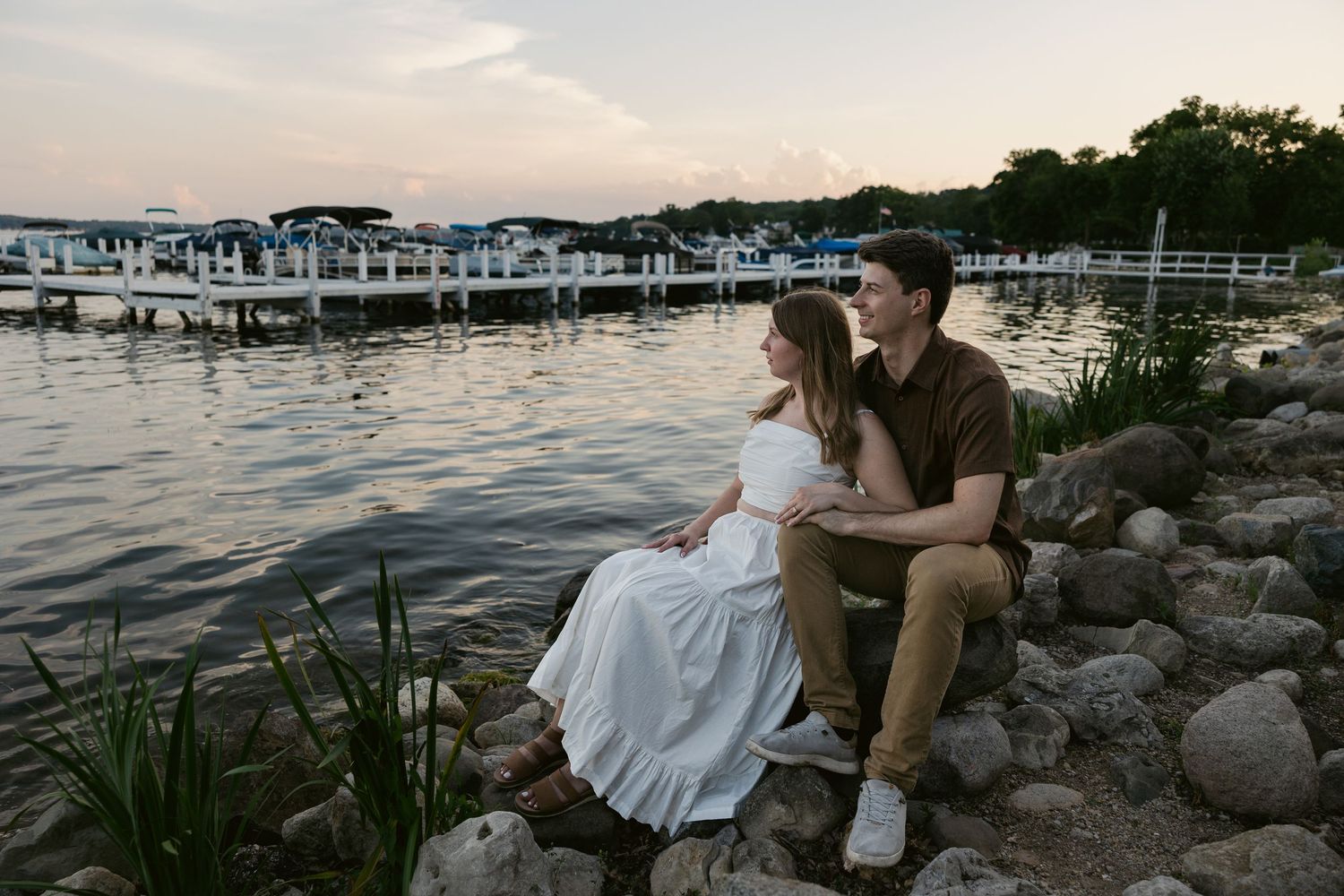 Couple sits on rocky shoreline with marina and boats visible in background.