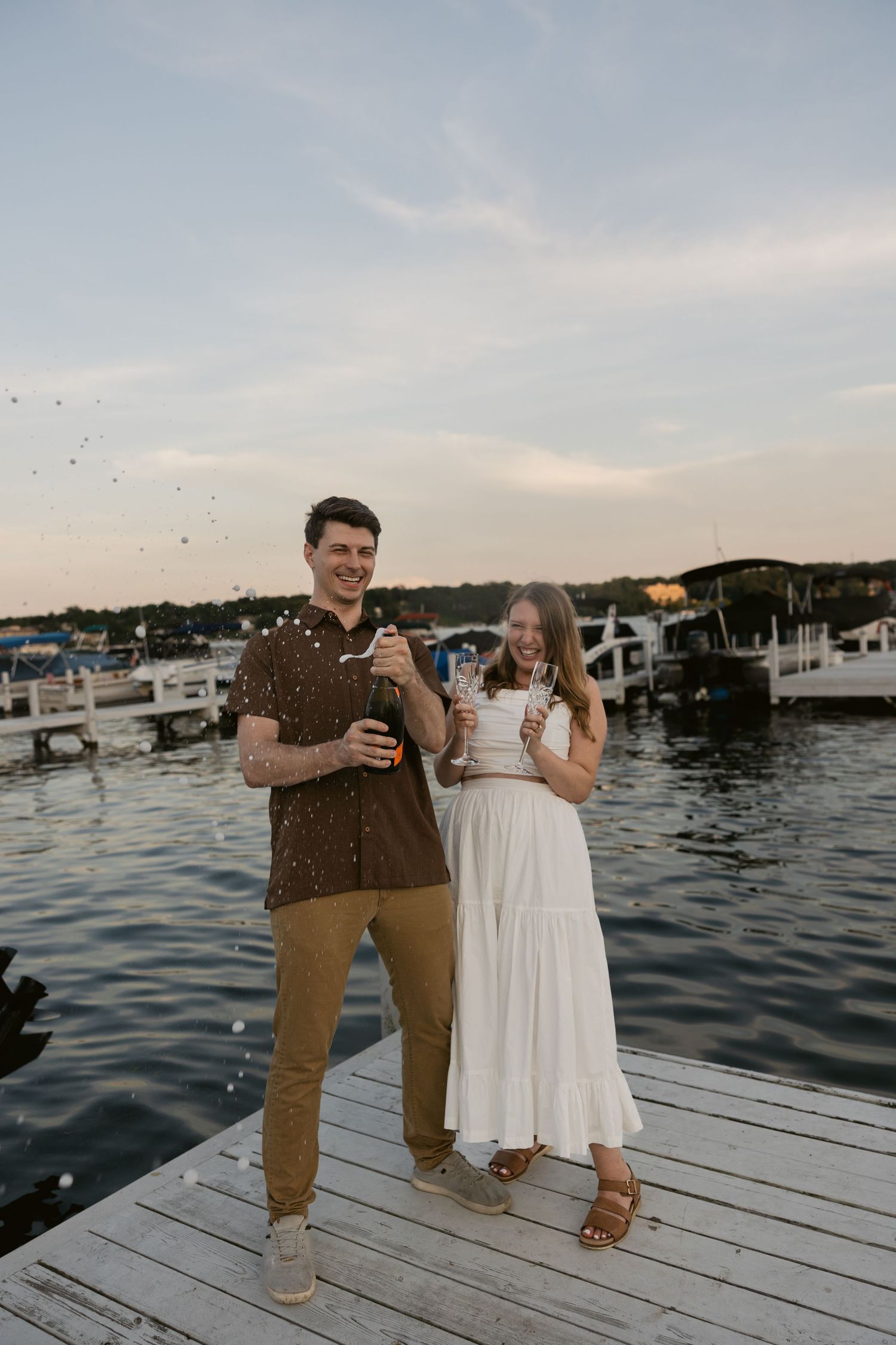 A couple celebrates on a waterfront dock with boats and marina in the background at dusk.