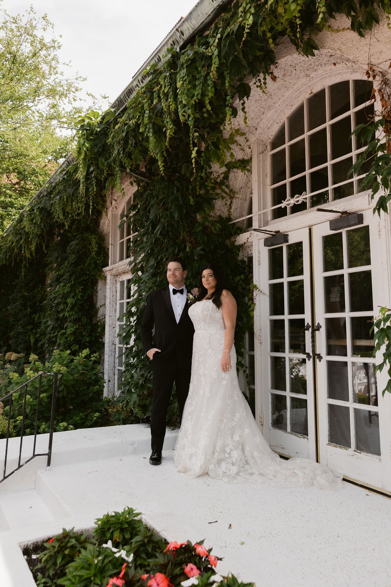 A newlywed couple poses against an ivy-covered building with elegant French doors and white trim.